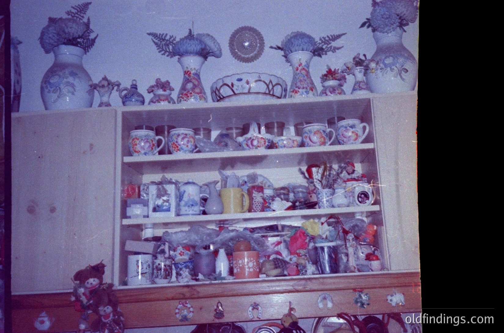 Vintage wooden shelf displaying handcrafted ceramic and glassware, likely Eastern European folk art from the 1960s–70s. Decorative motifs include floral patterns, geometric designs, and traditional symbols. Items include mugs, vases, plates, and figurines arranged on three shelves. Warm, earthy tones dominate with faded blue wall backdrop. Ideal for historical research on Soviet-era craftsmanship or interior design references.