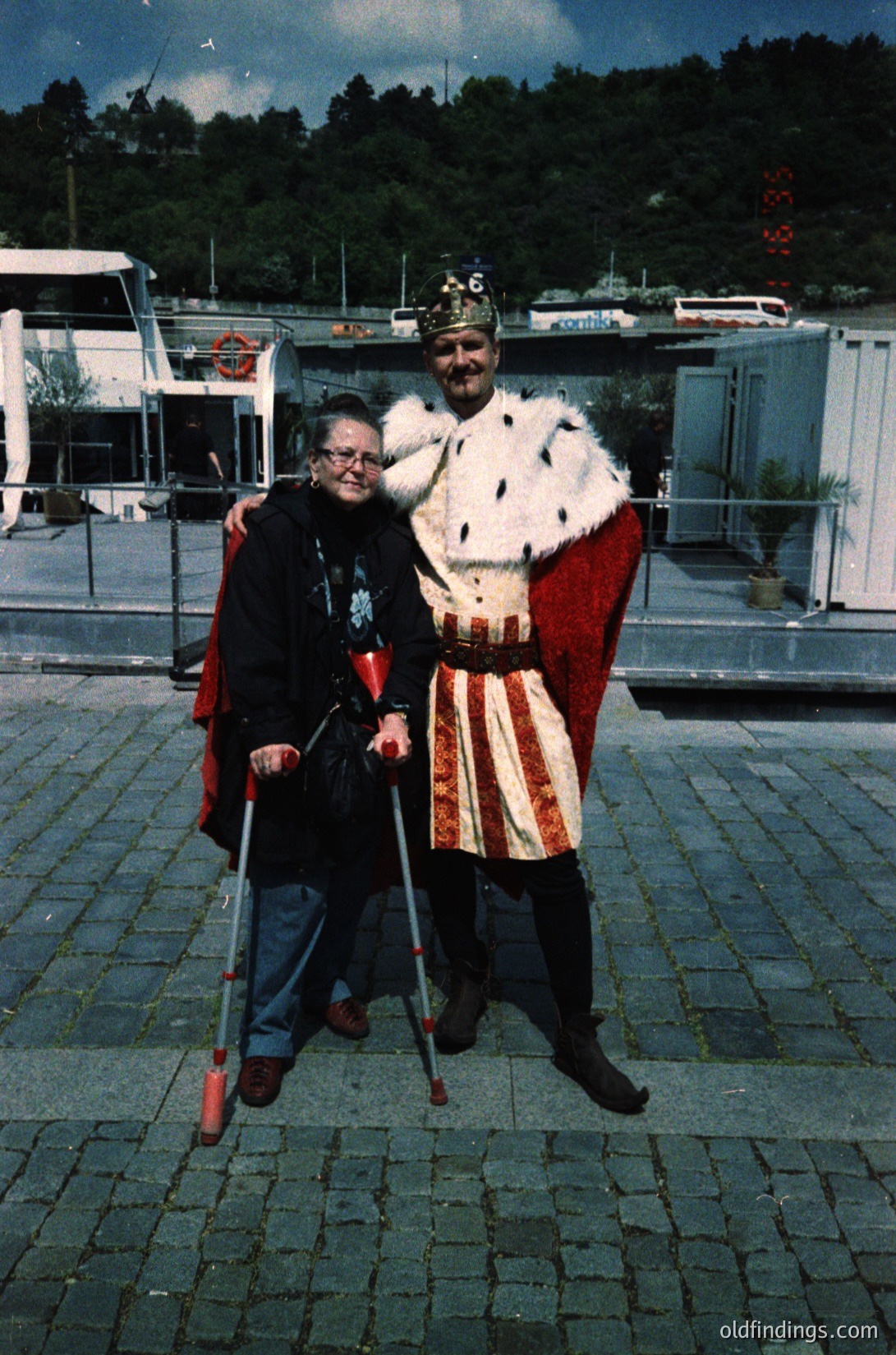 Vintage photo of two individuals in medieval-inspired attire on a cobblestone plaza, likely from the 1970s–1980s. The man wears a fur-lined cloak, crown, and striped tunic; the woman stands with crutches, dressed in a dark cloak. Mobile homes and greenery in the background suggest a festival or reenactment setting.