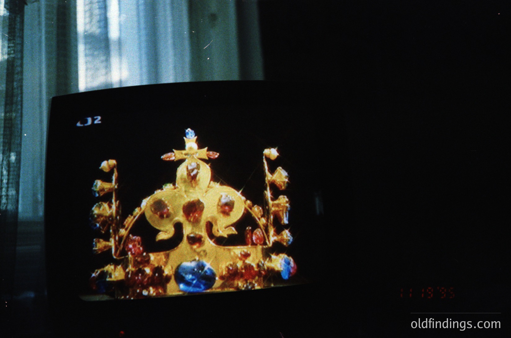 Vintage close-up of an ornate, gold-toned crown adorned with red and blue gemstones, likely a religious or ceremonial artifact. The intricate craftsmanship suggests Eastern Orthodox or Byzantine influence. Blurred background indicates indoor lighting, possibly from a museum or church setting.