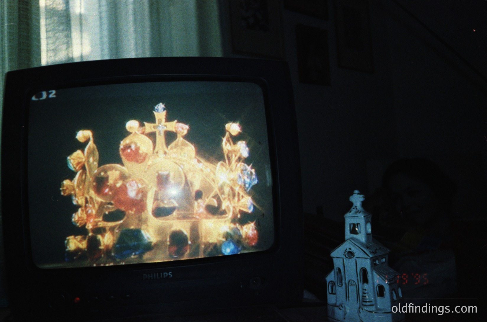 Vintage CRT TV displaying an ornate Orthodox religious icon, likely depicting a saint or Virgin Mary with haloed figures and floral motifs. Side table holds a small ceramic church model with a steeple and clock. Indoor setting suggests mid-20th century domestic decor.