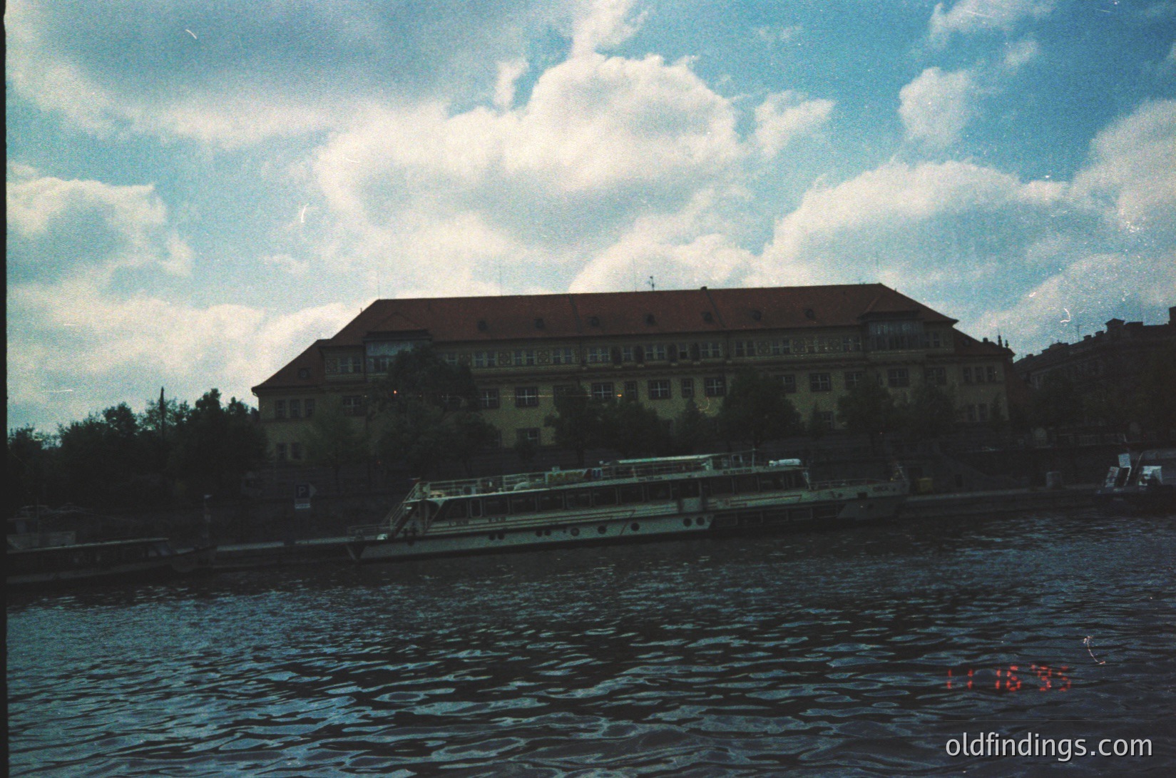 Large, multi-story building with flat roof and symmetrical windows, likely institutional or public architecture, positioned along a riverbank. A vintage passenger boat with open-air seating docked in foreground. Overcast sky with dramatic cloud formations. Timestamp suggests 1980s.