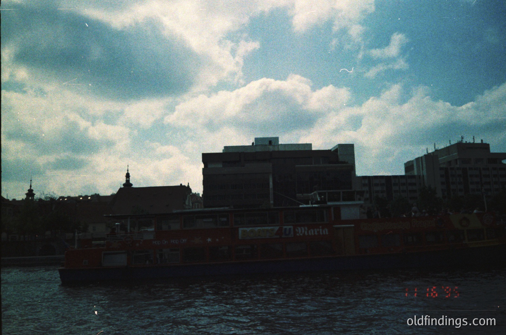 Vintage urban riverside scene with mid-20th century architecture. Prominent brick building with "Marina" signage, likely a hotel or restaurant, alongside a docked vessel. Distant skyline features a church spire and high-rise blocks under a dramatic sky. Date stamp reads 11/18/95.