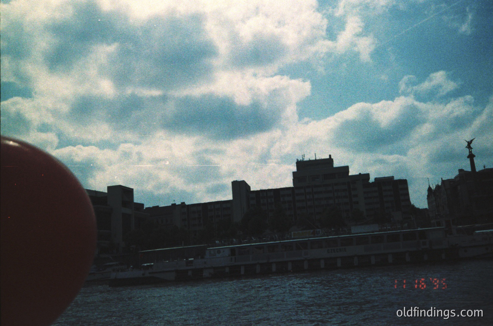 Vintage urban waterfront scene with mid-rise concrete buildings under dramatic cloud-lined sky. Bridge spans across dark water, timestamp 11:16:35 suggests 1980s-90s era. Industrial-era architecture with raw concrete textures.