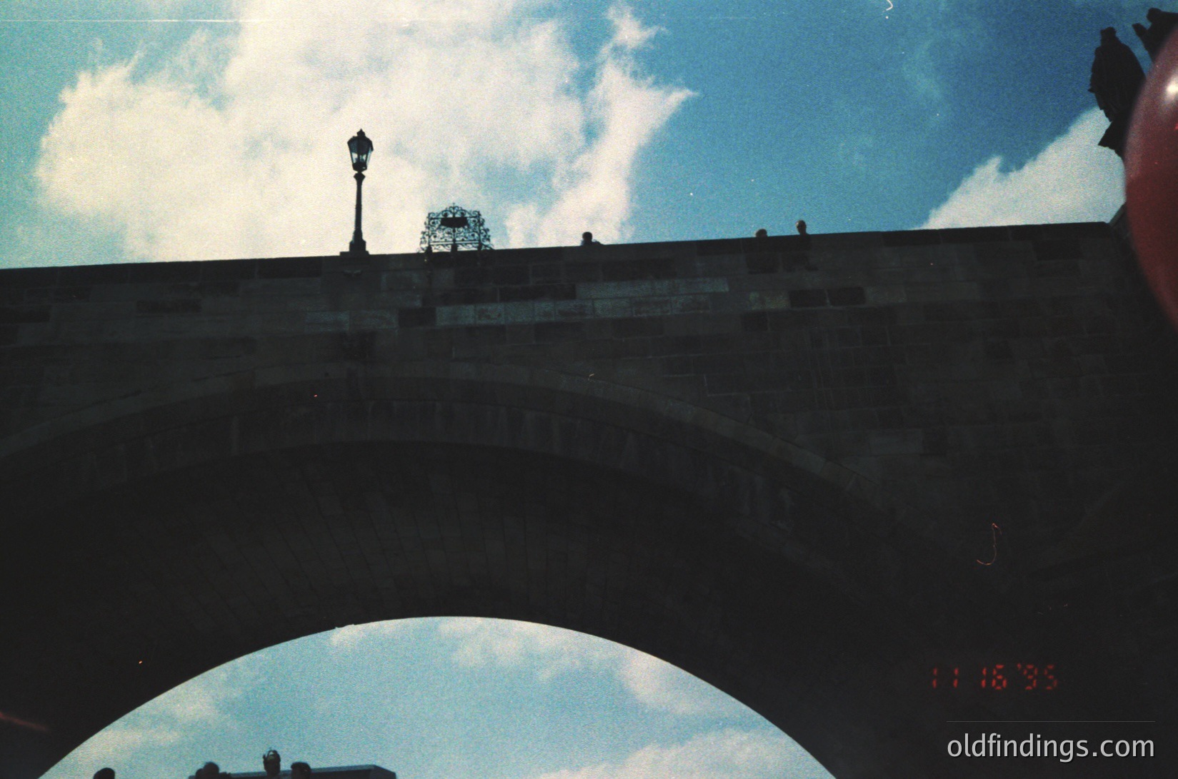 Vintage urban bridge arch with vintage street lamp and decorative wrought-iron structure atop. Reflections in wet pavement suggest recent rain. Time stamp "11:48:35" visible. Likely mid-20th century European cityscape.