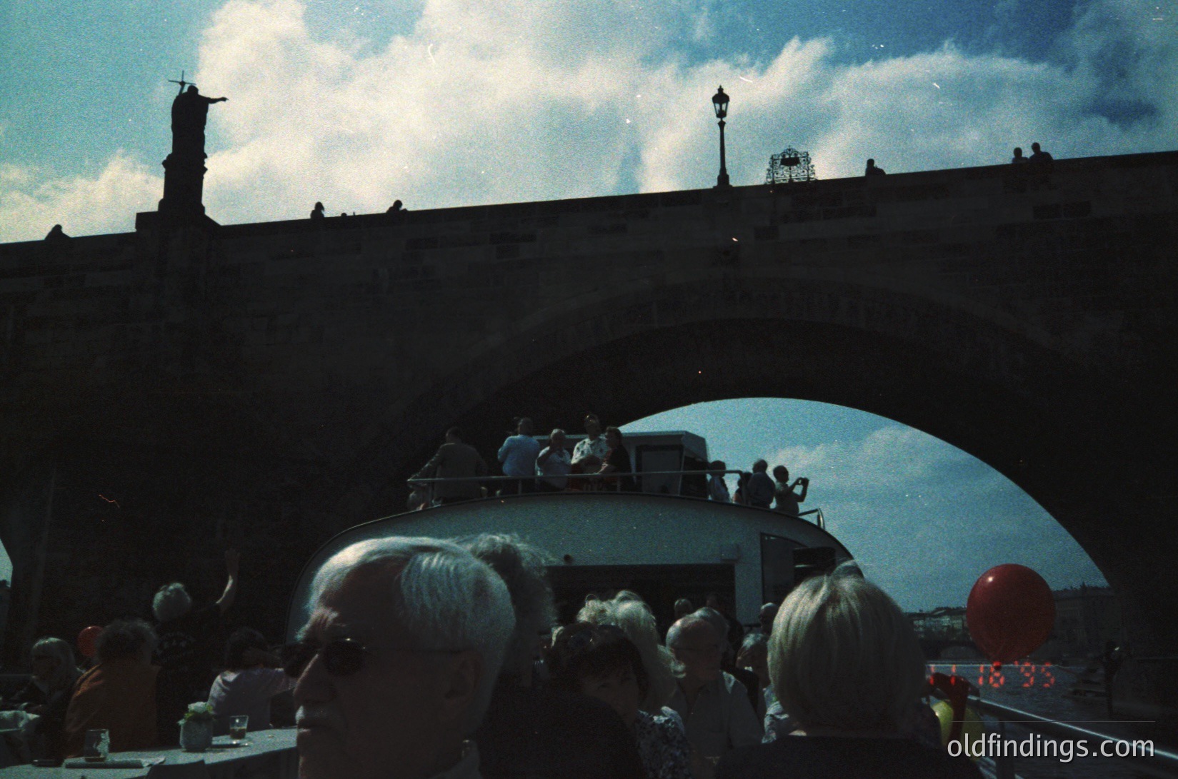 Vintage boat tour crowd on a river, likely Prague’s Vltava River, with Charles Bridge’s statues and lampposts in silhouette. Mid-20th century (1950s–1960s) attire and boat design suggest historical tourism. Crowded outdoor seating with a red balloon and clock reading 18:33.