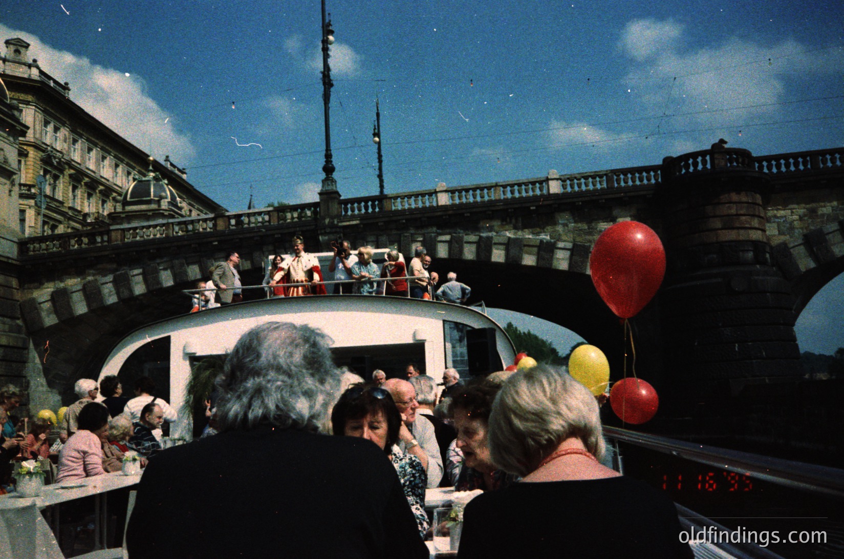 Vintage boat cruise with mid-20th century passengers enjoying a sunny day. Crowded deck features red/yellow balloons, retro clothing, and a clock displaying "11:30." Historic European cityscape with ornate bridges and classical architecture in background.