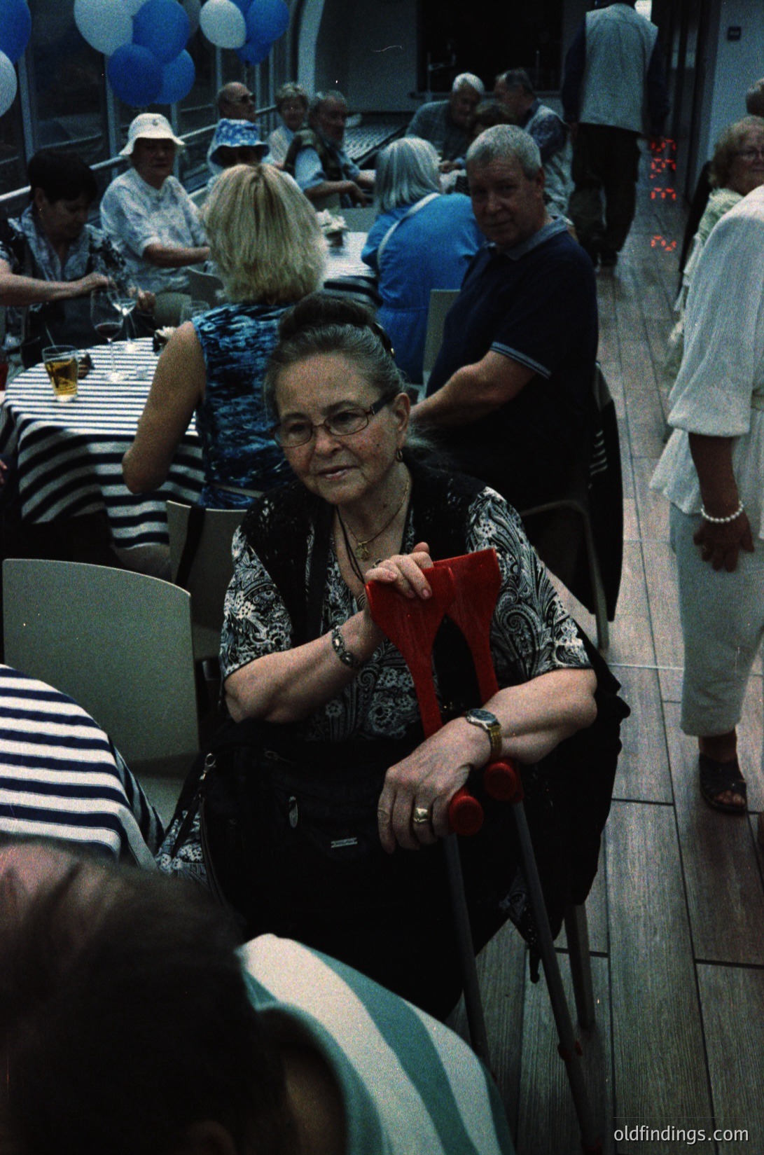 Vintage indoor gathering featuring a woman in patterned blouse and red scarf, seated at a long table surrounded by elderly guests. Decor includes striped tablecloths, balloons, and casual summer attire. Likely a community event or celebration, possibly or .