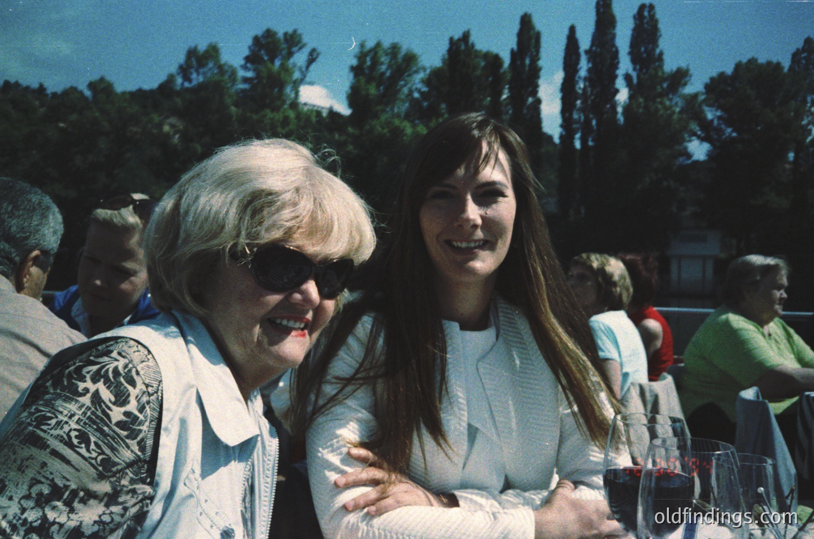 Two women share a joyful moment at an outdoor gathering, likely mid-1980s–1990s. The elder wears sunglasses and a patterned blazer over a light blouse; the younger, a white top with crossed arms. Glasses of red wine and blurred attendees suggest a relaxed social event.