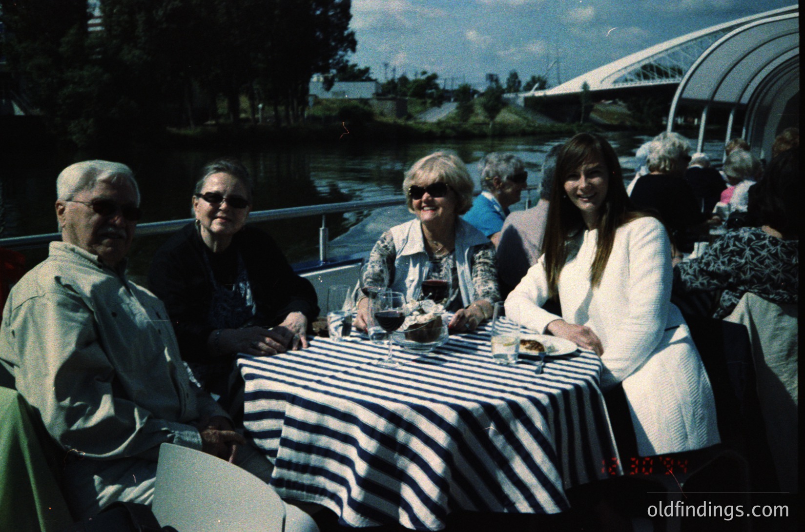 Four adults seated at a riverside café table, mid-1980s–90s. Men in short-sleeve dress shirts, women in patterned blouses and blazers. Striped tablecloth, wine glasses, and greenery in background. Likely European urban setting. éCulture