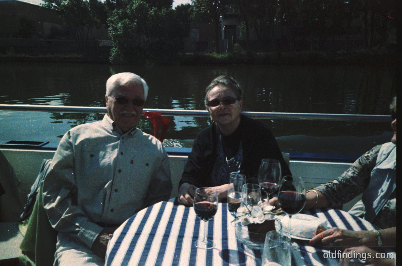 Vintage outdoor dining scene by a canal, featuring two elderly individuals seated at a striped tablecloth. Glasses of red wine and empty plates suggest a relaxed meal. Lighting and clothing hint at mid-20th century (1950s–1970s). Urban waterfront setting with greenery and boats in background.