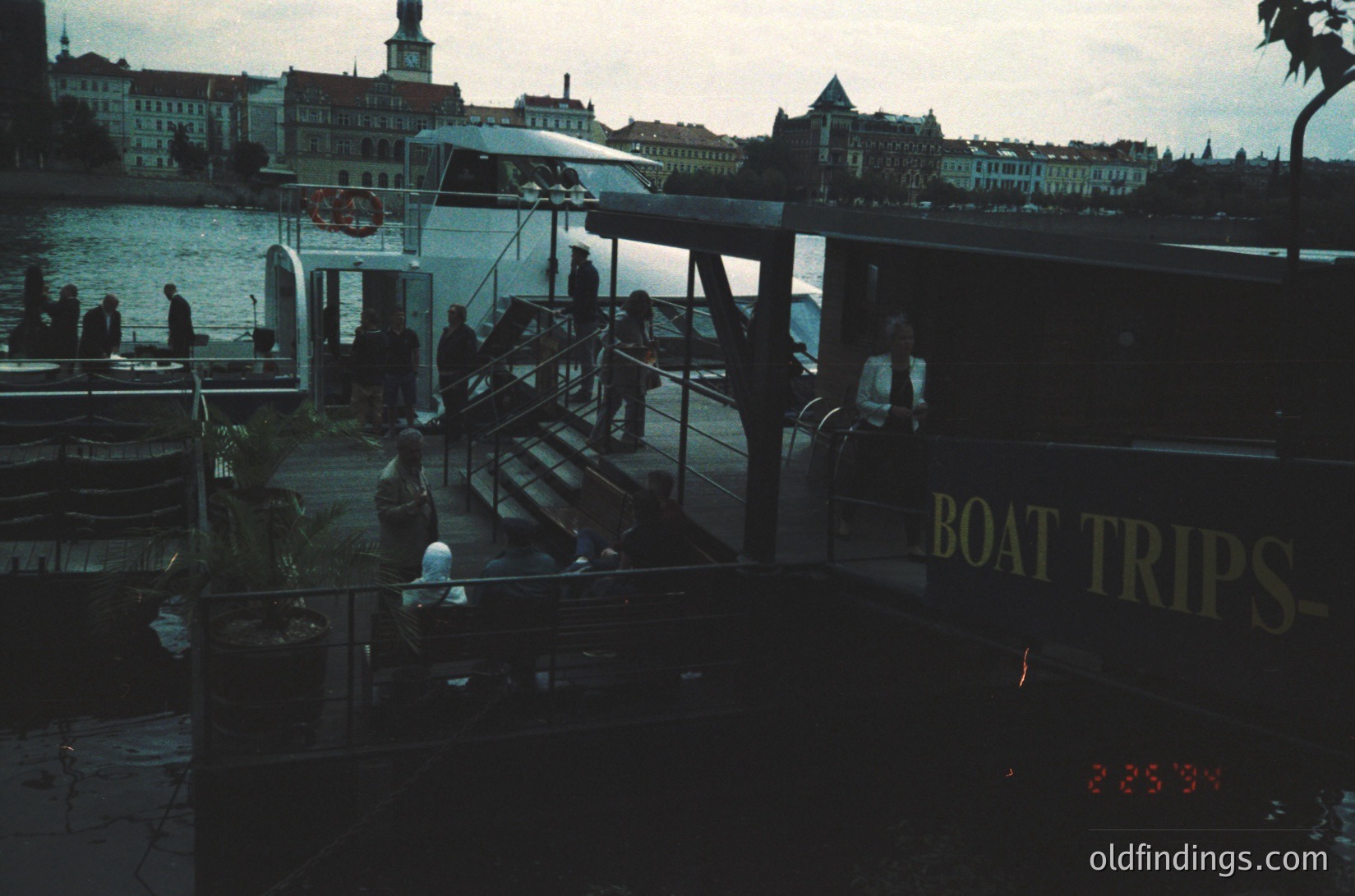 Vintage boat dock with "BOAT TRIPS" signage, likely Stockholm’s Gamla Stan district. Classic 1970s color sepia tone captures historic waterfront activity. Wooden pier, vintage ferry, and early evening crowd suggest leisure tourism.
