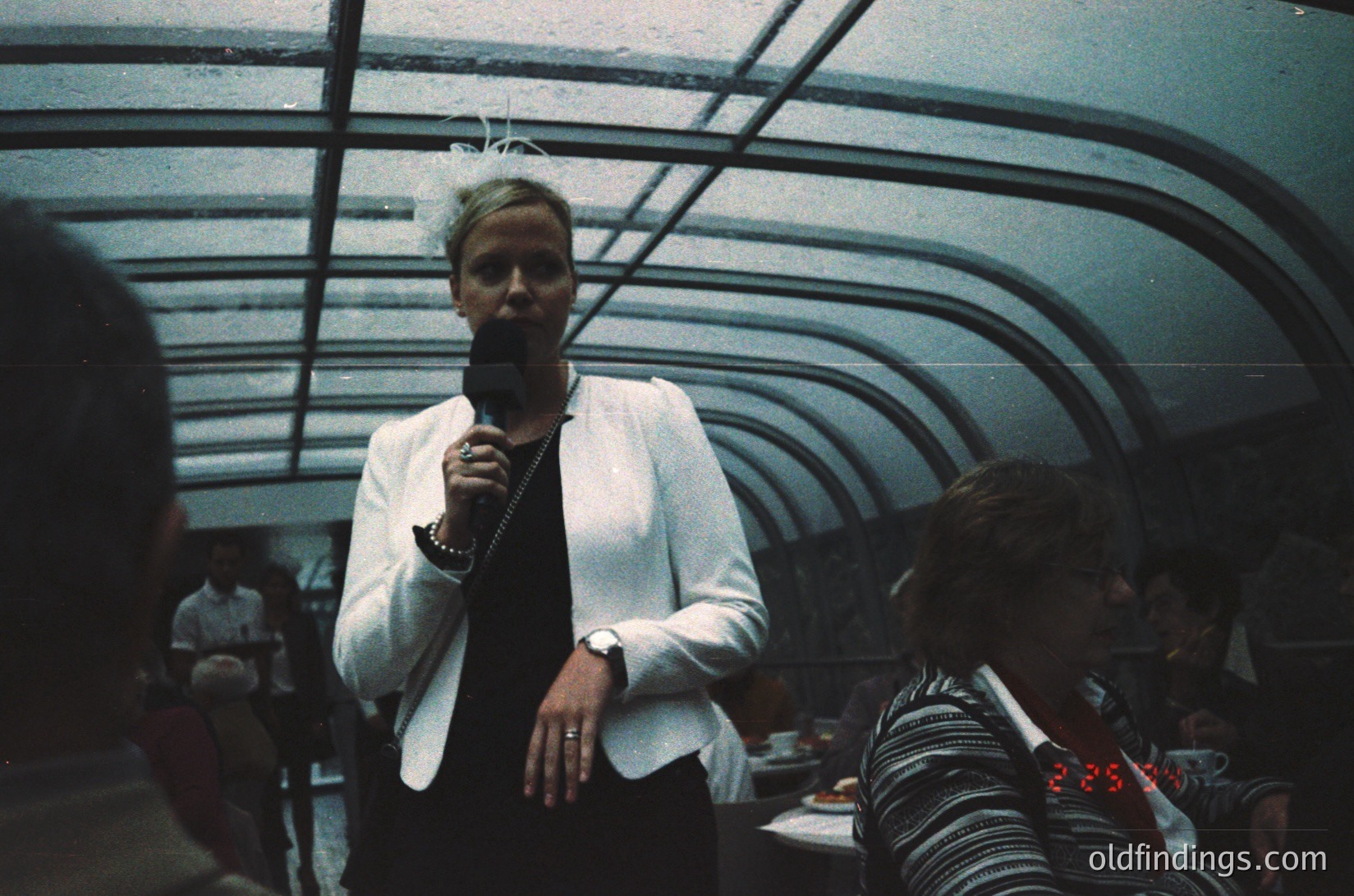 Vintage indoor concert shot featuring a performer holding a microphone in a dimly lit, curved-roof venue. Audience wears retro clothing; one person’s wristwatch displays red numbers. Likely 1970s–1980s live music scene.