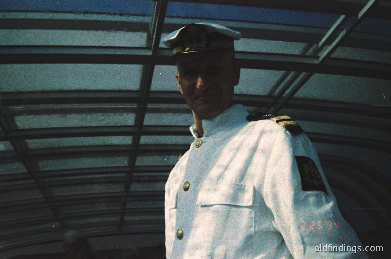Maritime officer in mid-20th-century naval uniform, standing under a metal bridge structure. White jacket with gold insignia, peaked cap, and rank patches. Visible date stamp "25.9.84" suggests 1984. Industrial or port setting.