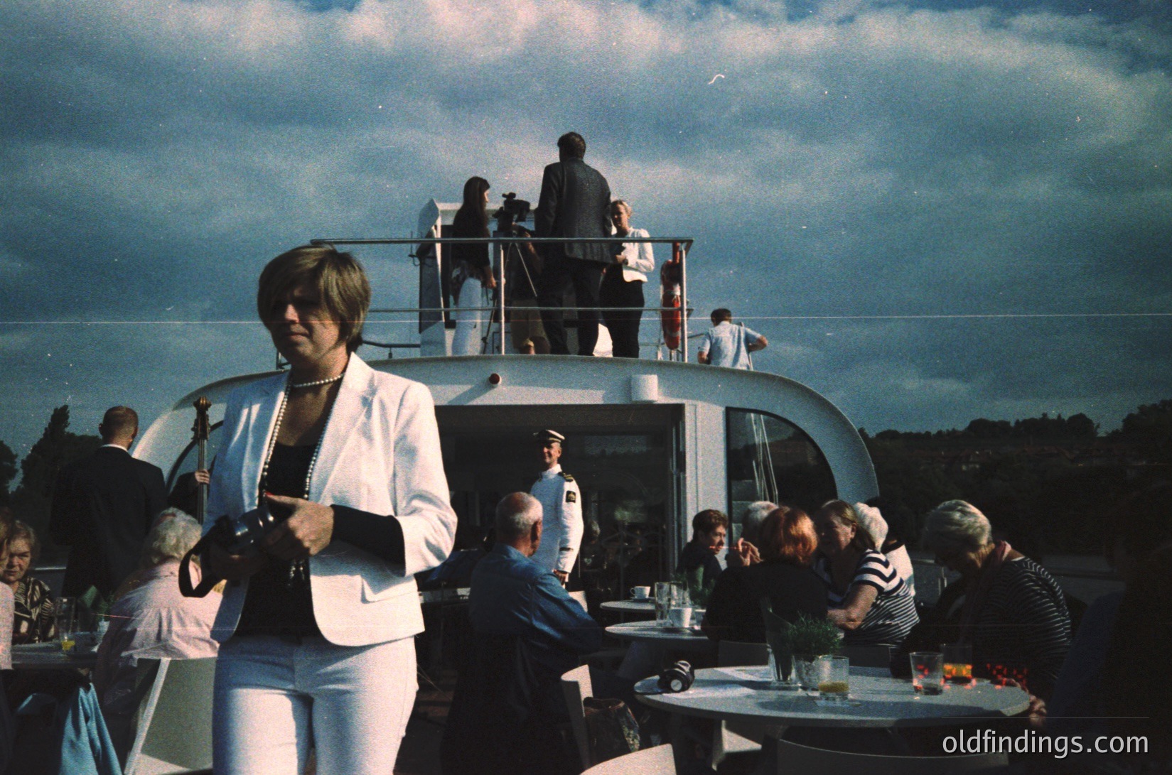 Vintage yacht deck party with mid-century attire. A woman in a white blazer holds a clipboard, overseeing guests seated at round tables with drinks and floral centerpieces. Upper deck guests stand near railings, overlooking a scenic, cloud-draped horizon. Likely 1960s–1970s European coastal setting.