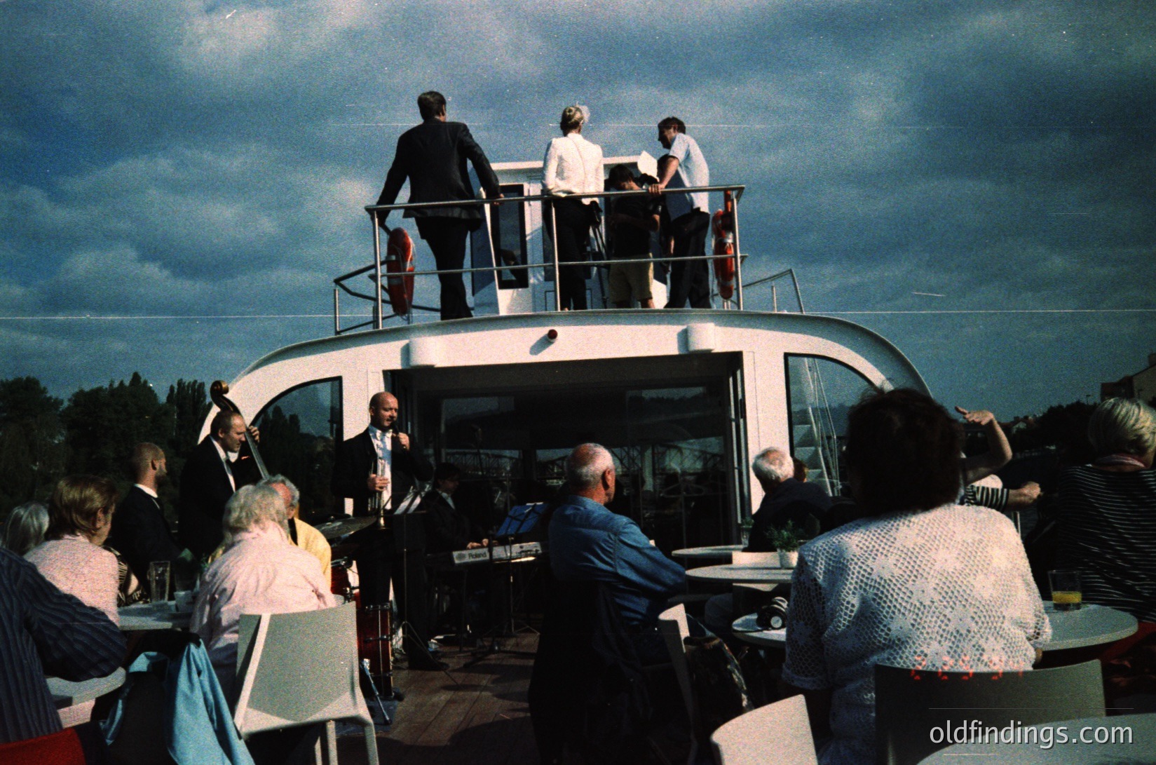 Vintage floating restaurant or observation deck with mid-century modern design, featuring a curved, white exterior and open-air seating. Six adults on upper deck in formal attire (1960s-70s style), while lower deck hosts a diverse group of patrons in casual summer wear. Overcast sky and greenery in background suggests a lakeside or riverside setting. Ideal for retro travel, hospitality, or architectural studies.