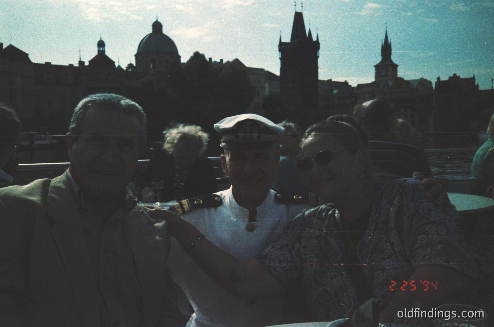 Vintage group photo in Prague’s Old Town, featuring 1960s-era attire (men in suits, women in patterned dresses). Iconic skyline of Prague Castle and Church of Our Lady before the Bridge Tower dominates background. Warm sepia tone suggests film photography.