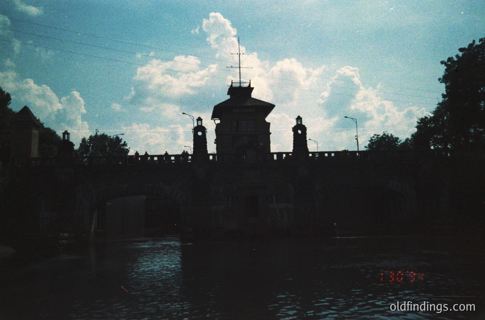 Silhouetted bridge tower with clock and decorative spires, framed by dramatic clouds and water reflection. Likely Eastern European urban architecture, mid-20th century.