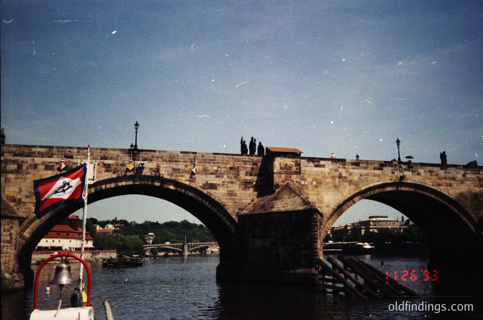 Historic stone bridge with Gothic arches spanning a river, featuring a flag with a red/blue emblem. Pedestrians and a boat with a bell in foreground. Likely Prague’s Charles Bridge (, , , , )