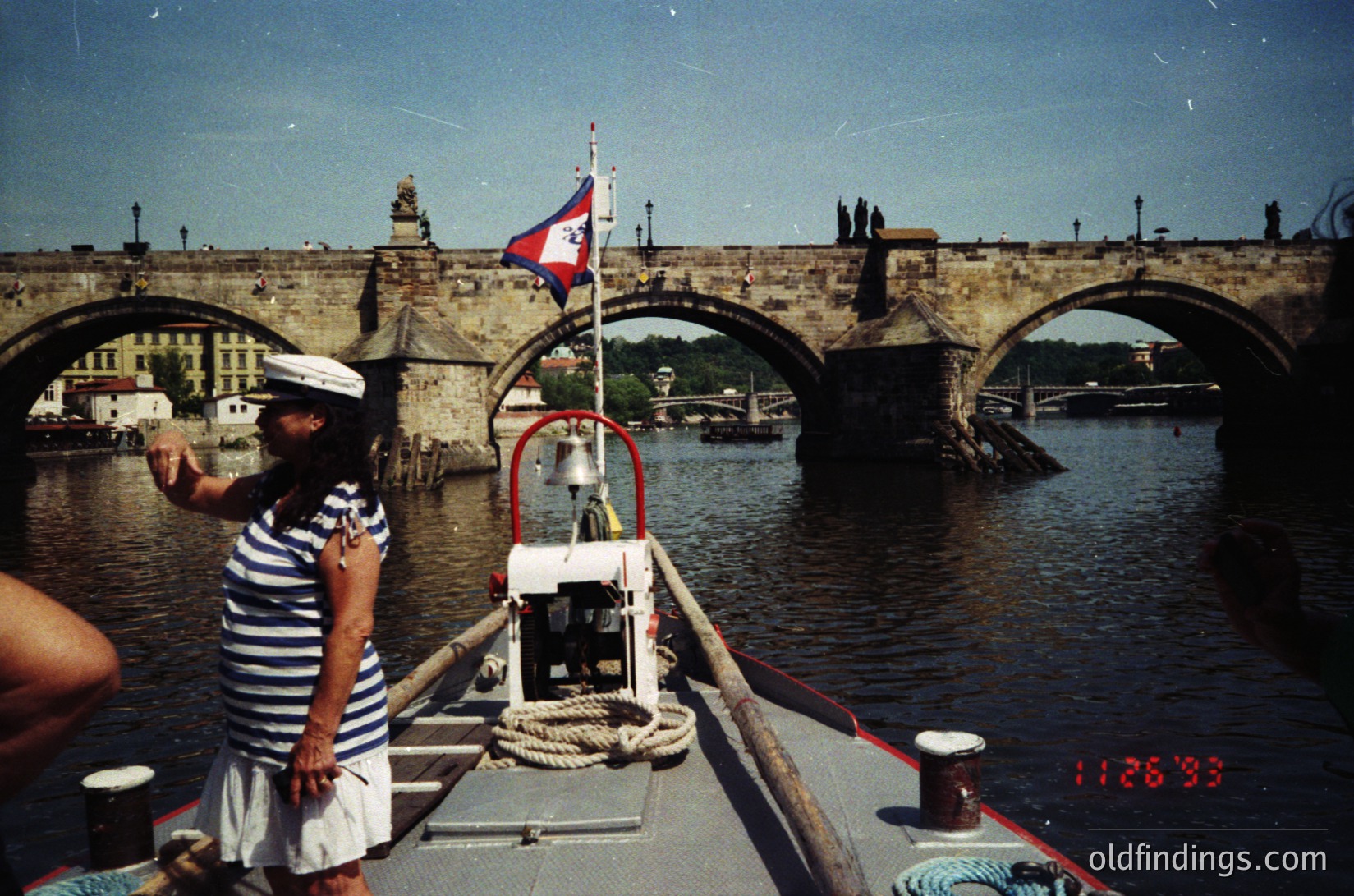 Vintage boat operator in striped uniform waves from Prague’s Charles Bridge, 1980s-90s. Gothic arches, historic flag, and river traffic highlight this iconic Czech landmark.