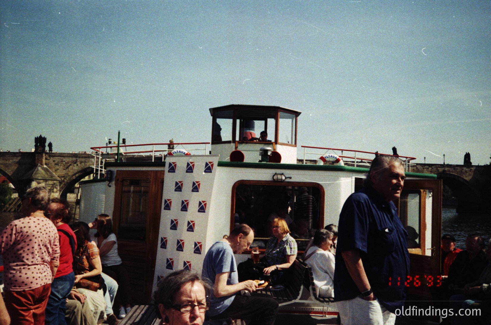 Vintage narrowboat with British flag-patterned trim, docked in a canal setting. Crowd of casually dressed passengers on deck, some seated, others standing. Clear vintage filter suggests 1980s–1990s era. Urban canal-side architecture visible in background.