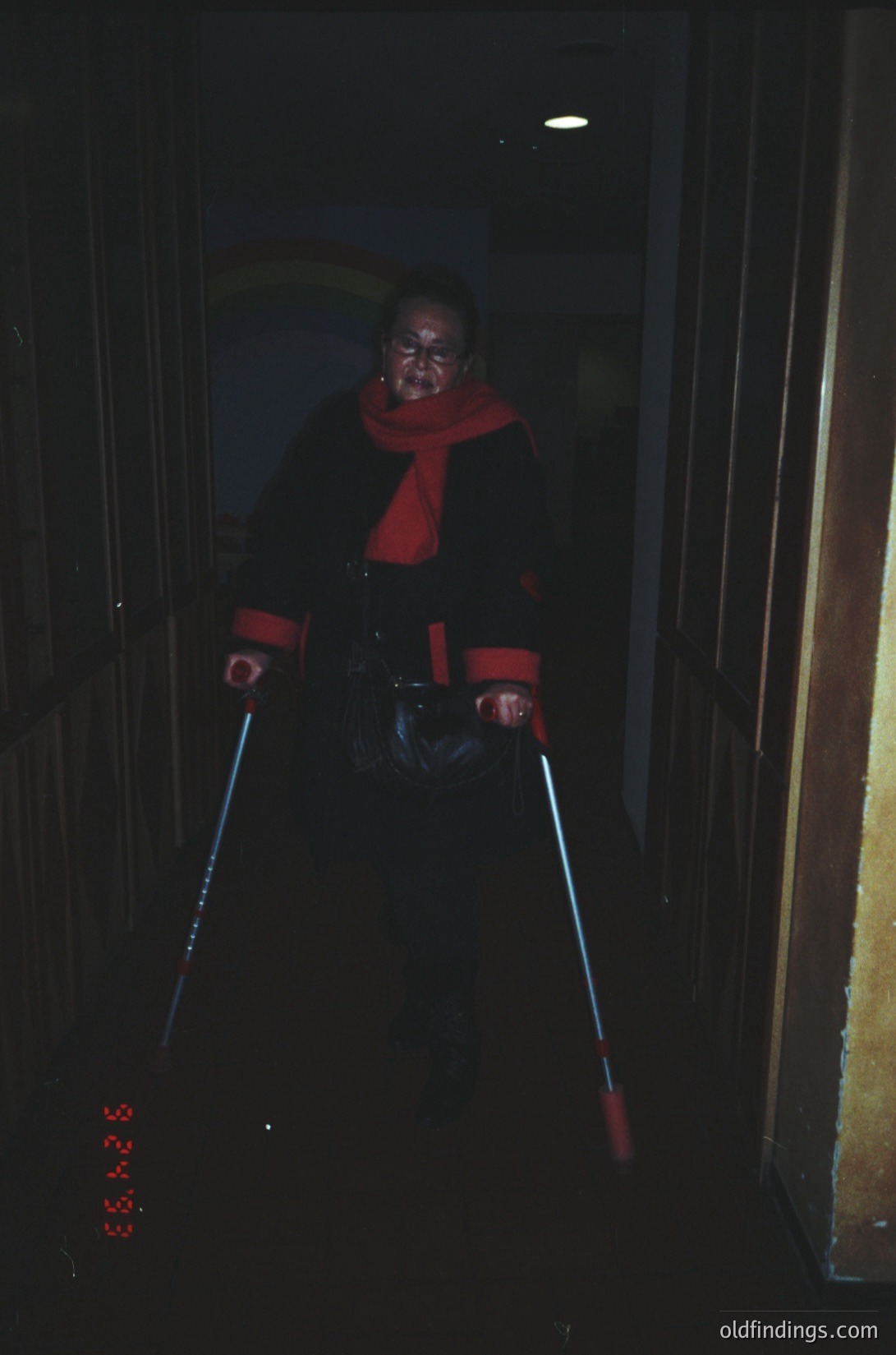 Vintage ski portrait in low-light indoor setting, likely a ski lodge or station. Person in red scarf and jacket, holding illuminated ski poles with neon tips, suggesting 1970s–1980s winter sports culture. Warm-toned film grain enhances nostalgic atmosphere.