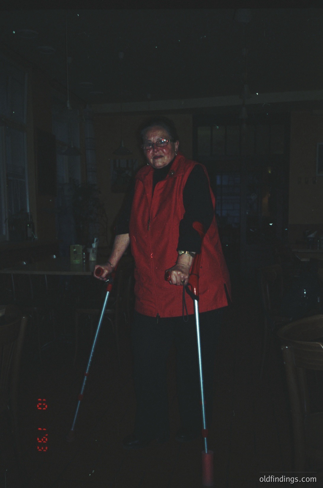 Vintage black-and-white photo of elderly individual in a red vest, holding cane-like sticks, indoors near kitchen appliances. Face painted red, suggesting a festive or theatrical event. Likely 1960s–1970s era, possibly Halloween or carnival.