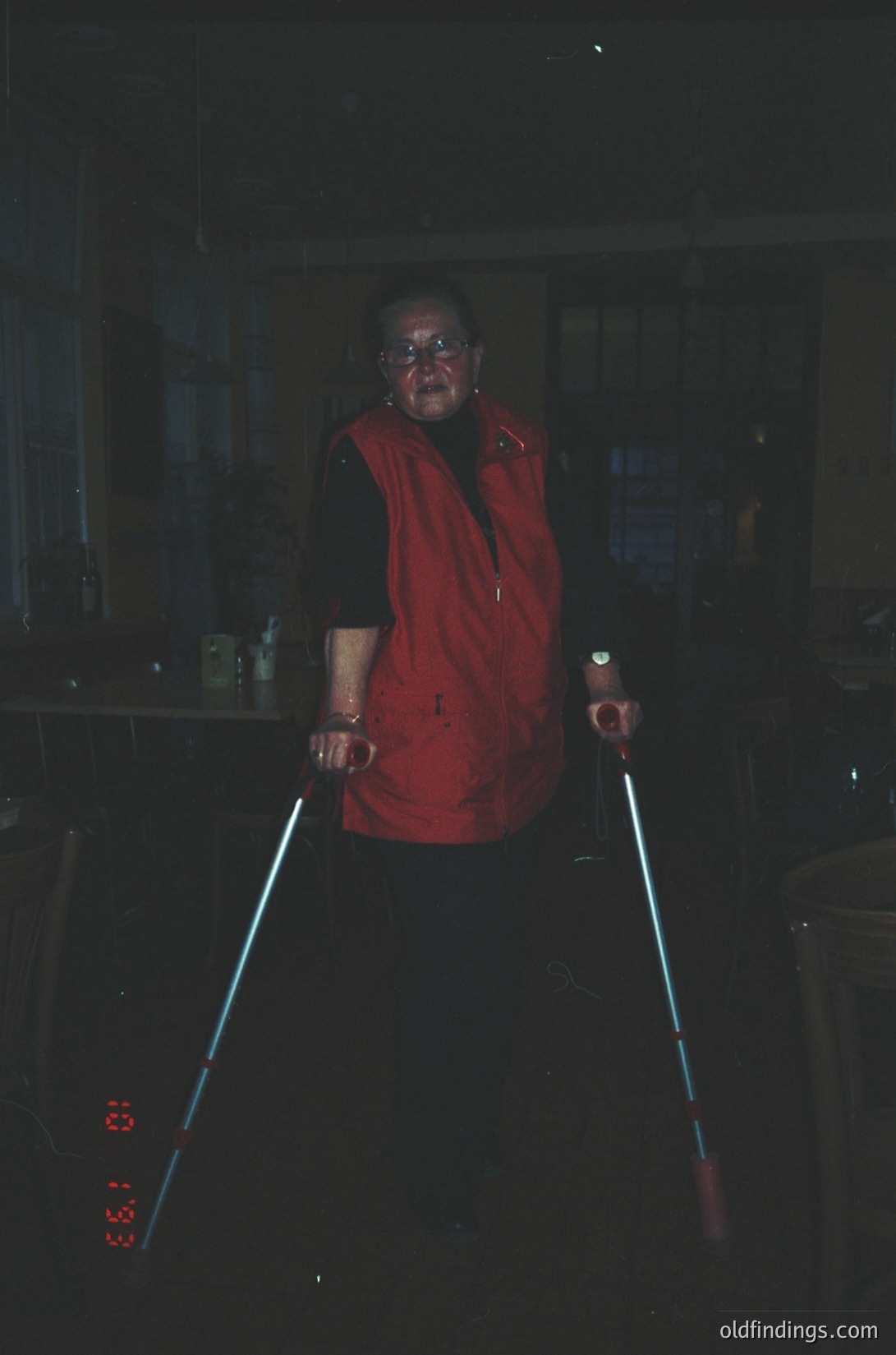 Indoor portrait of an elderly woman using illuminated walking sticks in low-light, likely a 1970s–1980s kitchen. She wears a red blouse, round glasses, and a choker necklace. The scene suggests limited visibility, possibly due to aging or lighting conditions.