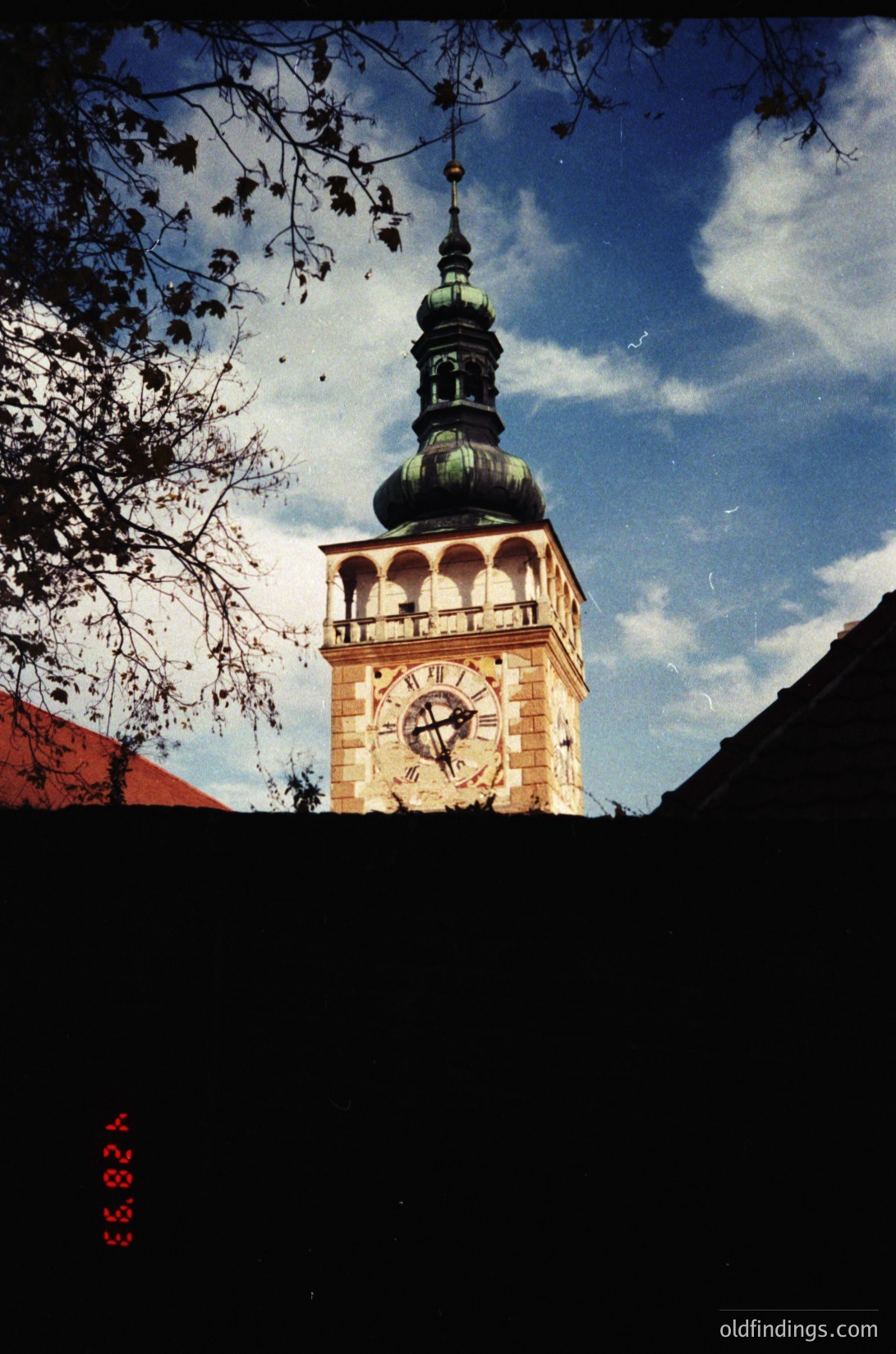 Historic clock tower with Baroque-style architecture, featuring a green-domed spire and ornate clock face. Leaning tree branches frame the upper portion, suggesting autumn or winter. Likely European, mid-20th century urban setting.