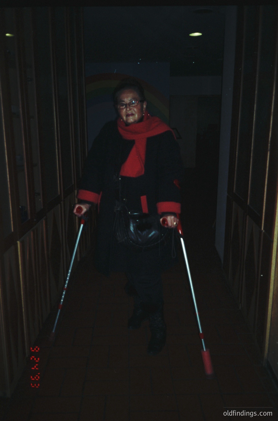Portrait of an elderly individual descending stairs using illuminated canes, likely for mobility assistance. Dark coat with red scarf and sunglasses. Indoor setting with vertical metal railings and dim lighting. Style suggests mid-to-late 20th century.