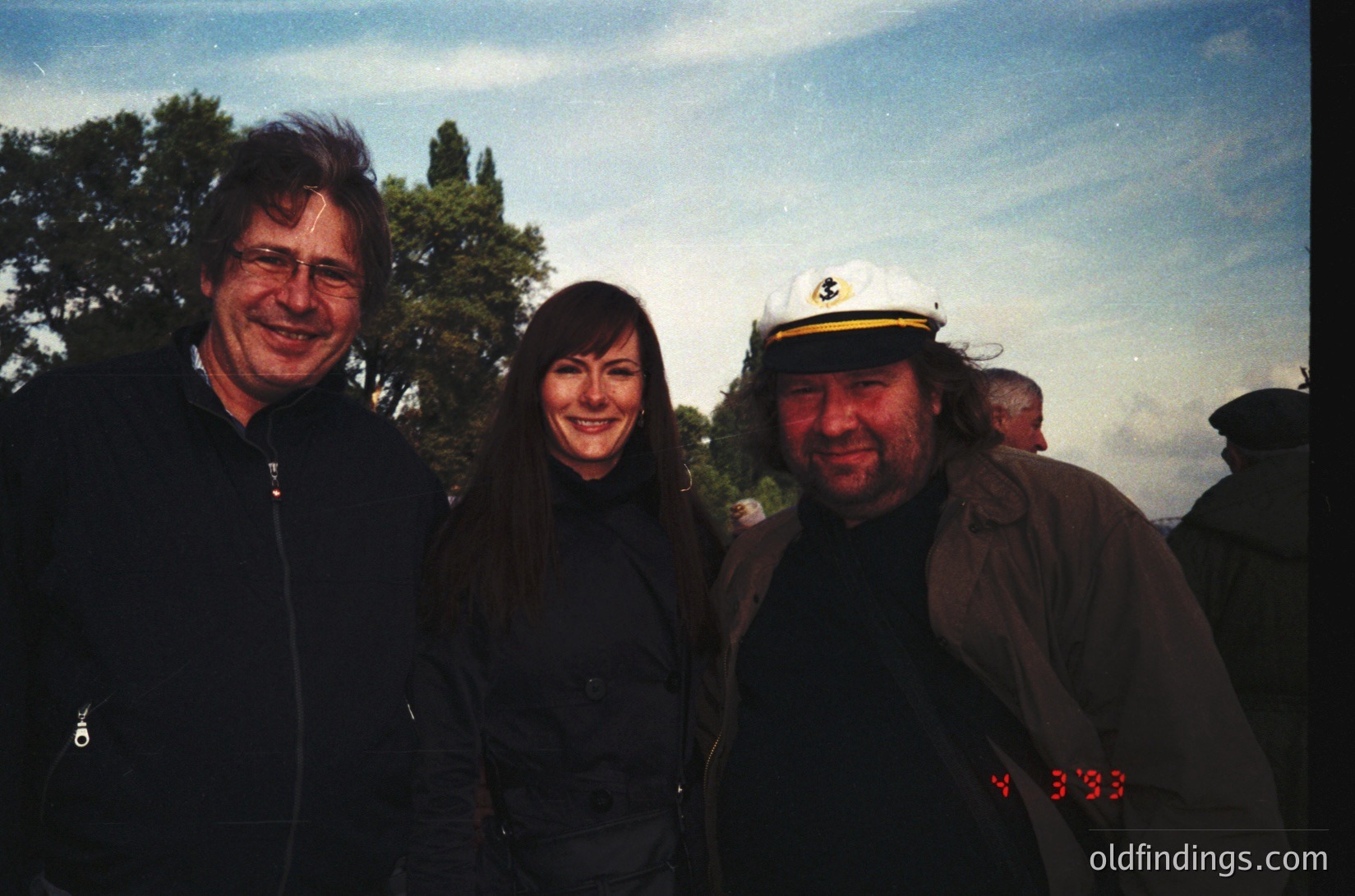 Three individuals pose outdoors in a vintage-style photo, likely late 20th century. The man on the left wears a dark jacket, the woman in the center a black coat, and the man on the right a sailor-style cap with a yellow emblem. Trees and a blurred background suggest a park or garden setting. Digital timestamp reads "4:33."