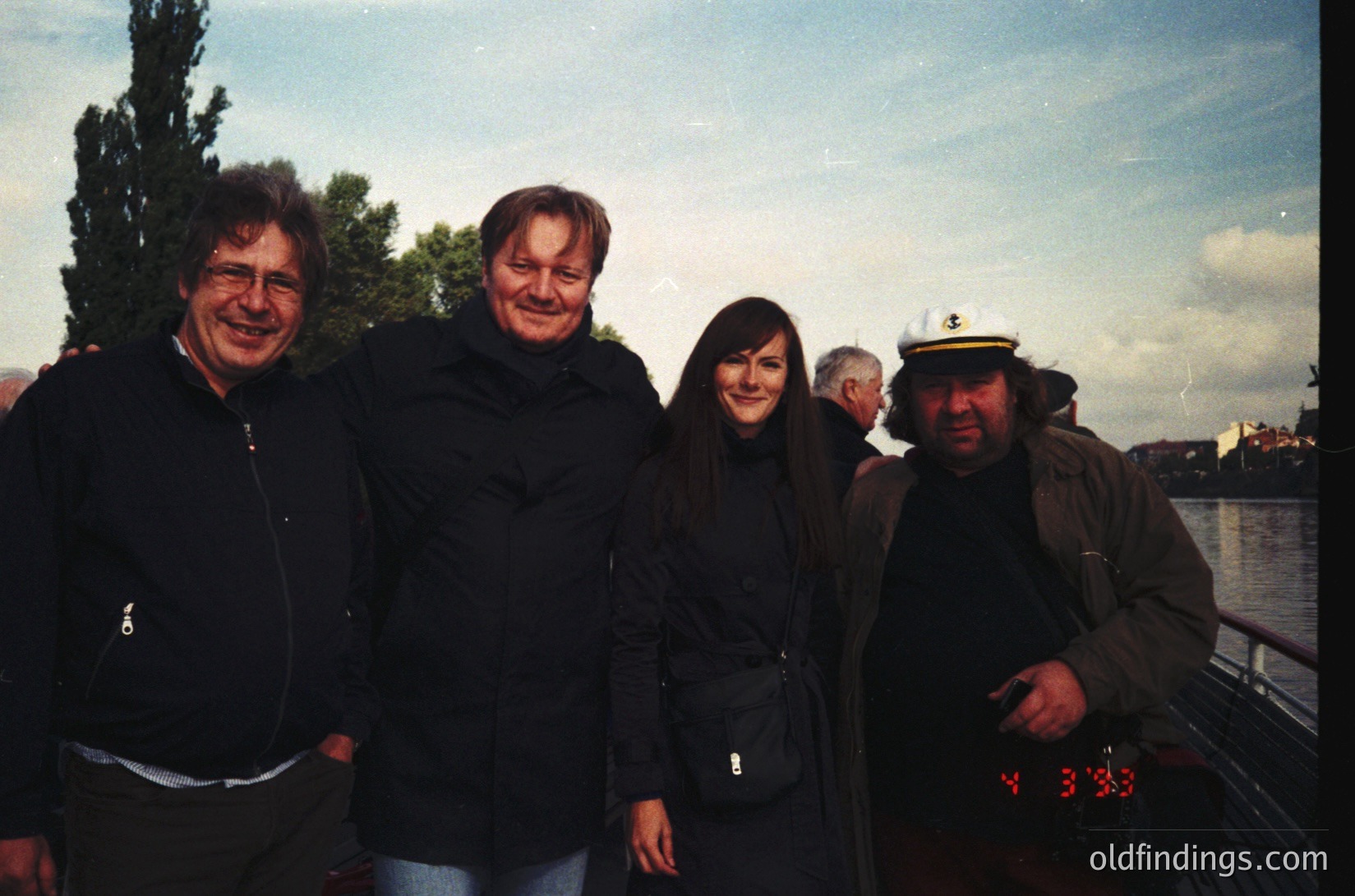 Four individuals pose outdoors near a waterfront at dusk, likely in a European city. The man on the right wears a vintage captain’s hat and a digital clock displaying "23:35." Casual attire suggests a relaxed social gathering. Urban skyline and bridge in background indicate a coastal or riverside location.