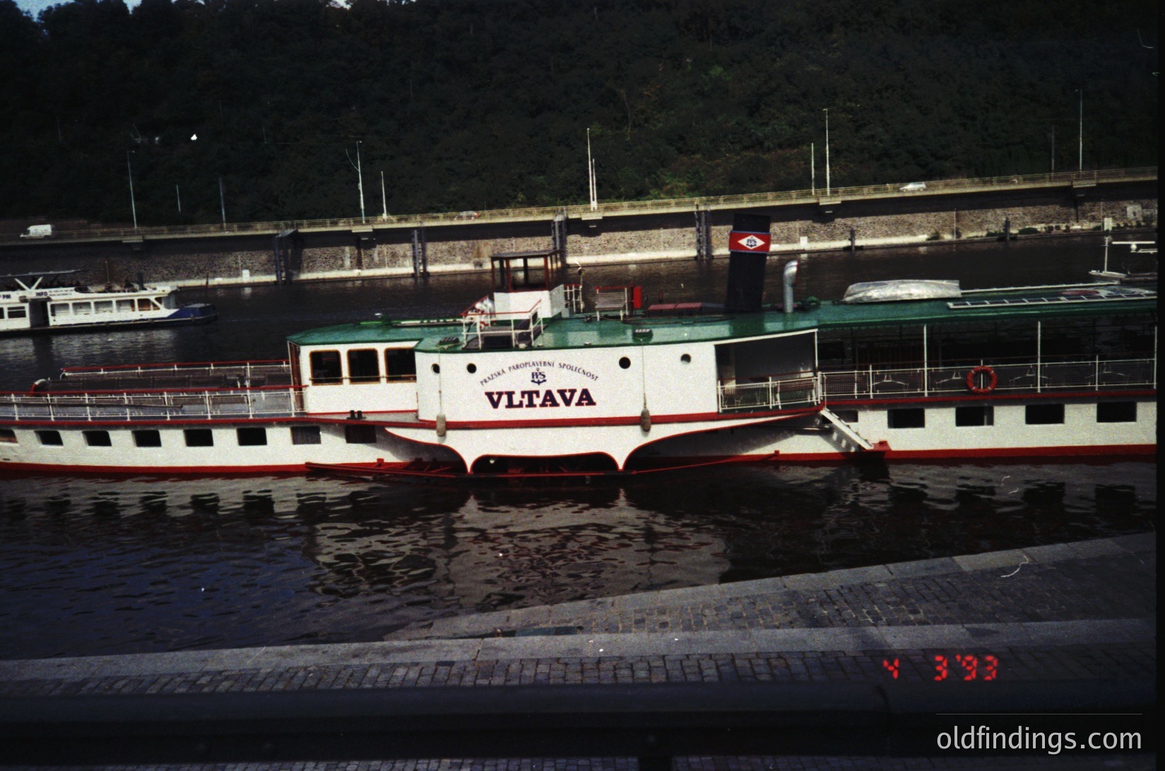 Vintage riverboat "Vltava" docked at Prague’s historic waterfront, featuring classic 1960s-70s passenger ferry design with red-and-white hull. Stone embankment and industrial-era bridge in background.