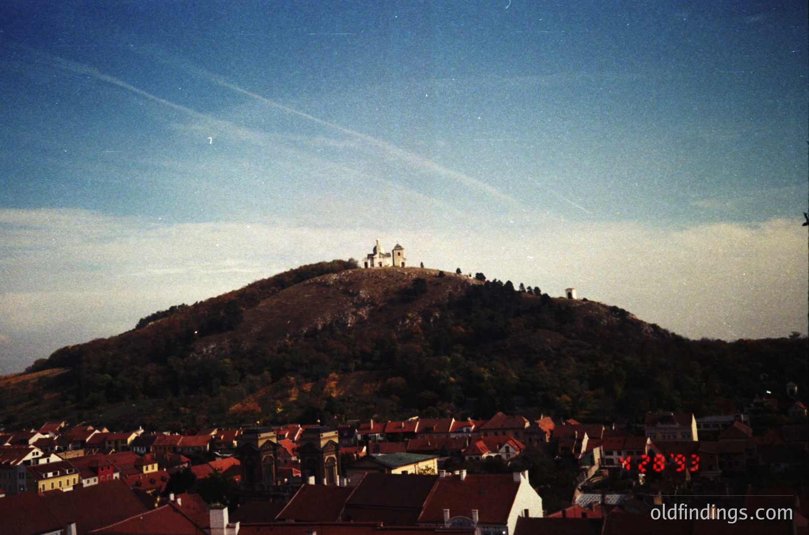 Vintage aerial view of a hilltop fortress atop a forested hill, surrounded by a densely built European town. The structure features a central tower with smaller buildings clustered around it. Foreground shows red-tiled rooftops and a neon sign reading "1983" in the lower right. Likely Eastern European, 1980s.