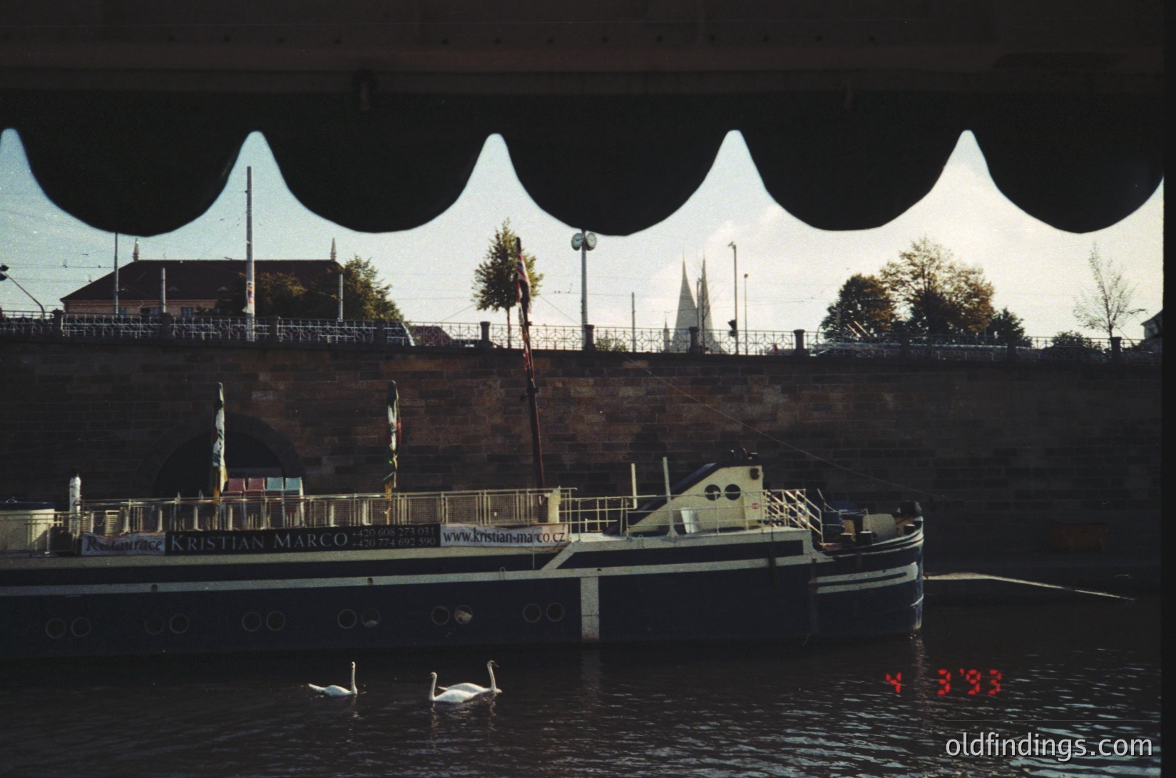 Vintage riverboat "Kristian Marco" docked under a modern, wave-like canopy bridge. Two swans glide near the vessel’s bow. Urban skyline with spires and greenery in background. Likely European riverfront, 1990s–2000s.