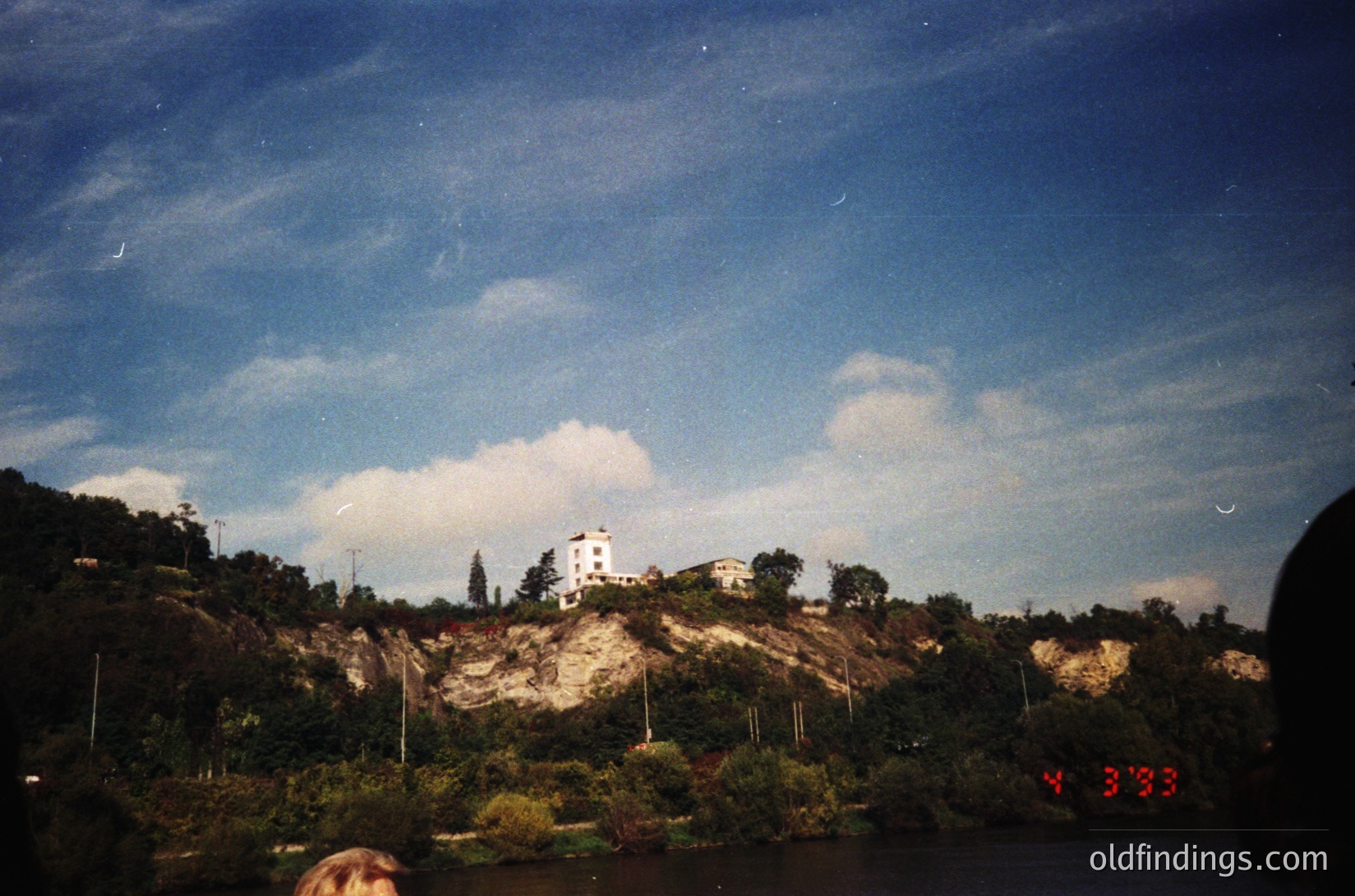 Vintage Polaroid-style shot of a coastal lighthouse perched on rocky terrain, surrounded by dense greenery. The timestamp "Y 3'33" suggests 1973. Dramatic sky with scattered clouds and a crescent moon.
