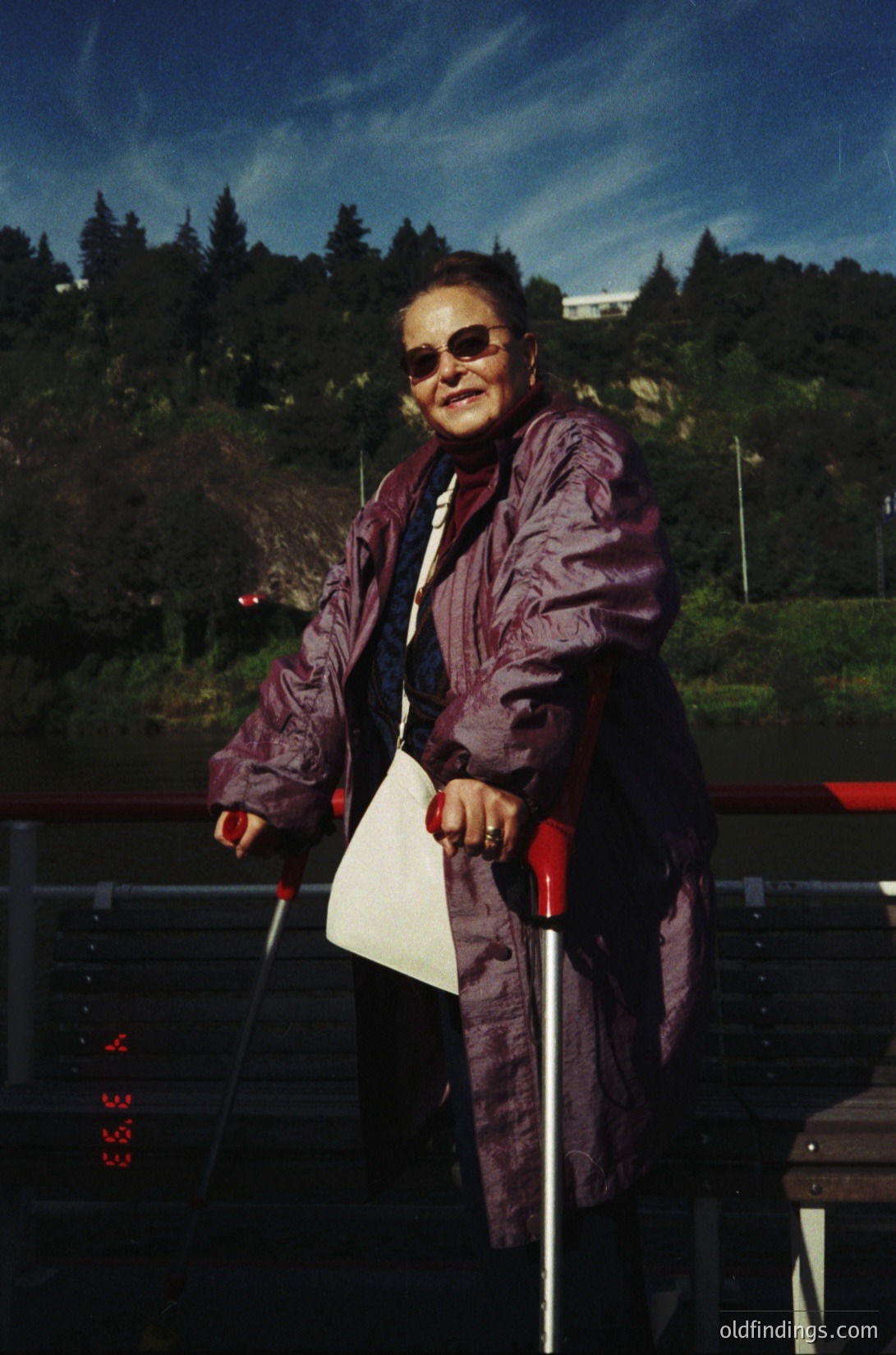 Portrait of an individual in a maroon coat and sunglasses, leaning on a cane with a white paper in hand. Outdoor setting with forested background and modern urban infrastructure (e.g., digital clock display: "3:33"). Likely 1980s–1990s, urban/suburban area.
