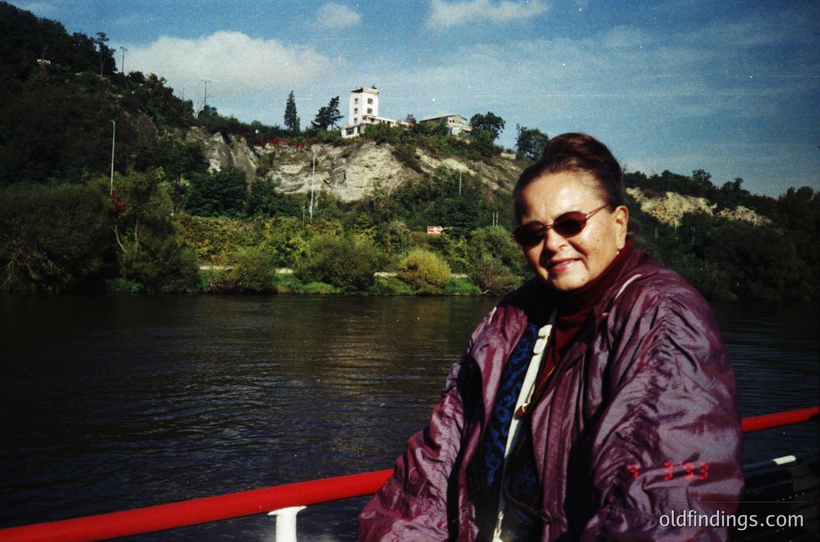 Woman in maroon jacket poses by a riverbank, wearing sunglasses and a lanyard. Rocky hillside with sparse vegetation and a white building in background. Likely 1980s-1990s outdoor portrait.