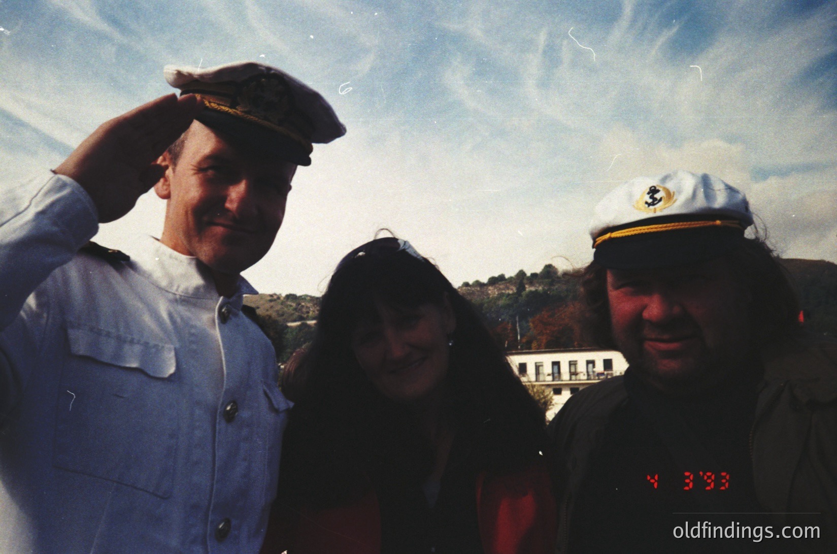 Three individuals pose outdoors in vintage attire, likely 1990s. Left: man in white naval-style cap and jacket. Center: woman in dark scarf and jacket. Right: man in white cap with insignia, wearing a red "1993" wristband. Background shows a hillside with a white building and clear sky.