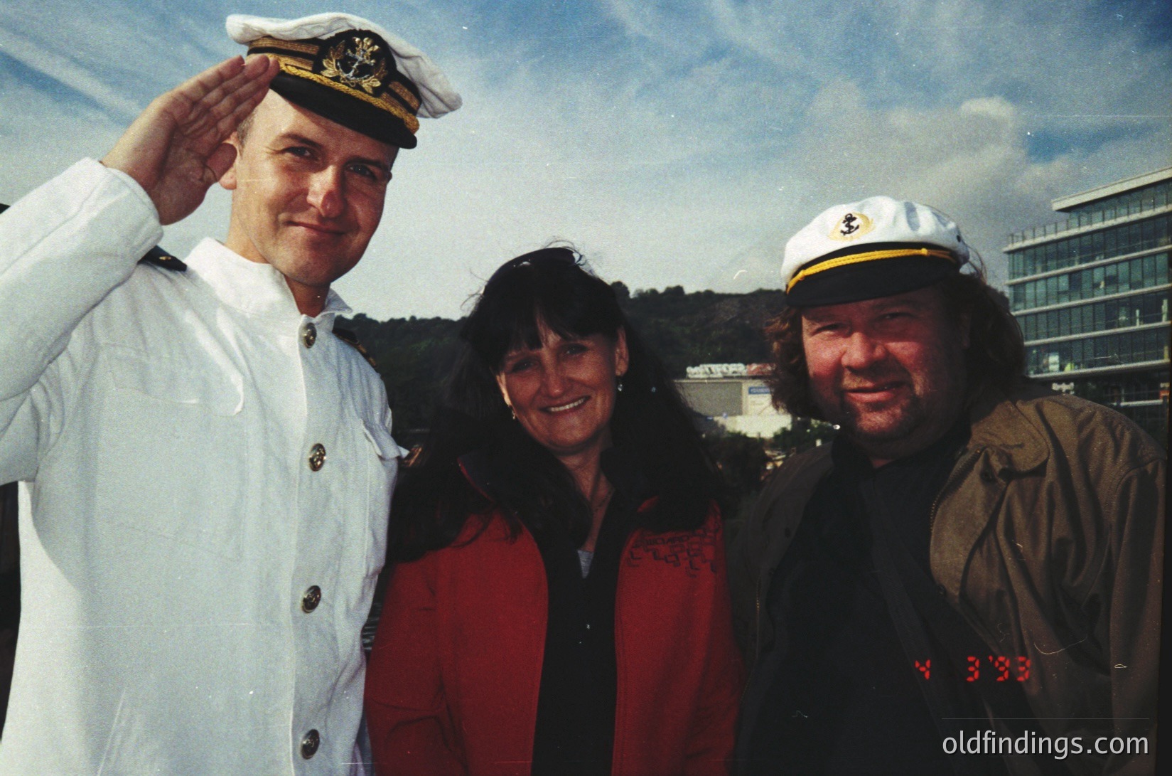 Three individuals pose outdoors in formal naval and casual attire, likely mid-1990s. The man on the left wears a white naval officer’s cap and uniform with gold insignia, while the woman in red stands beside him. The man on the right dons a sailor’s cap and a brown jacket with visible "333" embroidery. Urban background suggests a coastal cityscape.