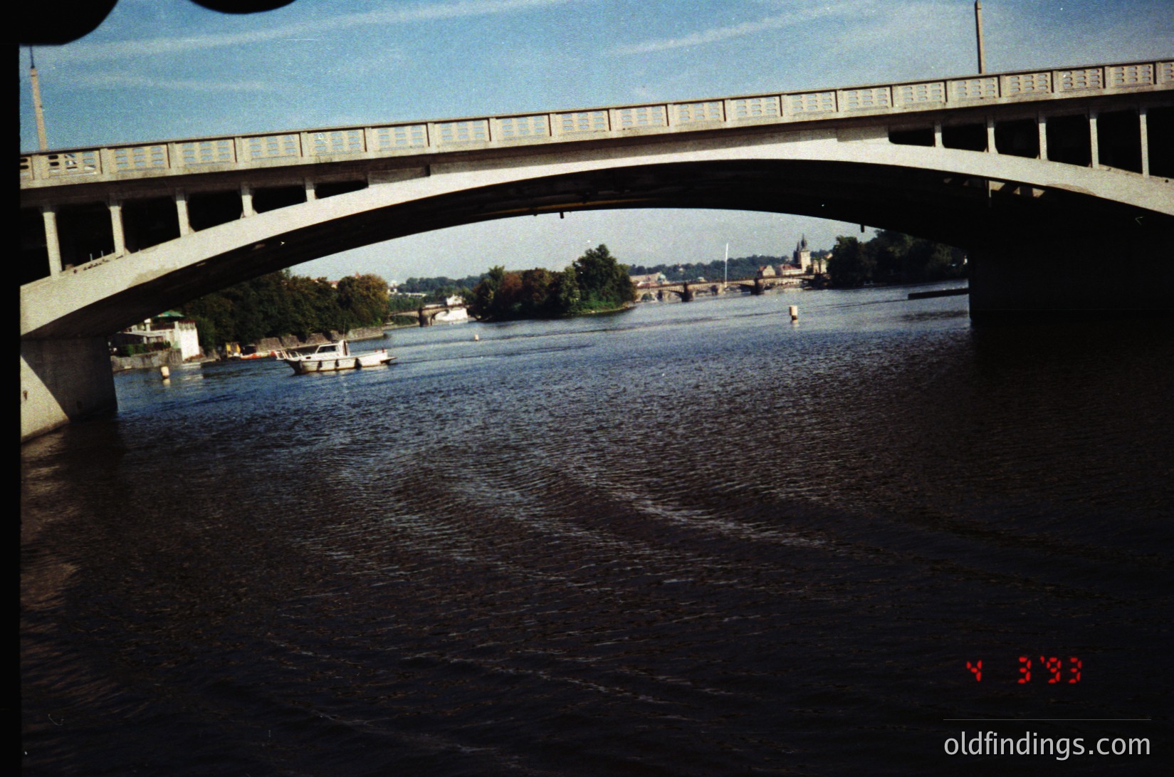 Vintage archival shot of a concrete bridge spanning a river, framed by its own arch. Mid-20th century urban landscape with boats on calm waters, distant industrial buildings, and greenery. Likely Eastern European due to architectural style.
