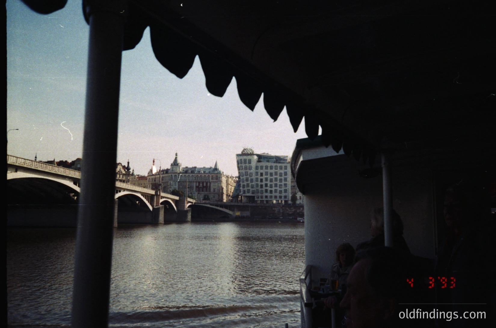 Vintage shot of a river scene framed by a boat’s scalloped edge, likely the Danube. Historic stone bridge and mid-century modernist buildings in background suggest Budapest, Hungary. Warm sepia tone and grain indicate 1960s–1970s era. Ideal for urban history, travel, or nostalgic design references.