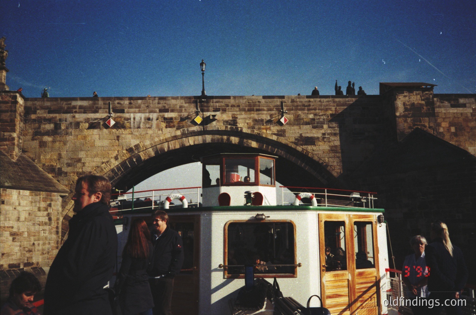 Historic boat tour beneath Prague Castle’s stone archway, 1990s. Wooden and glass-covered vessel with passengers enjoying views. Gothic architecture and medieval stonework frame the scene. Crowds gather on bridge and boat, capturing tourist activity.
