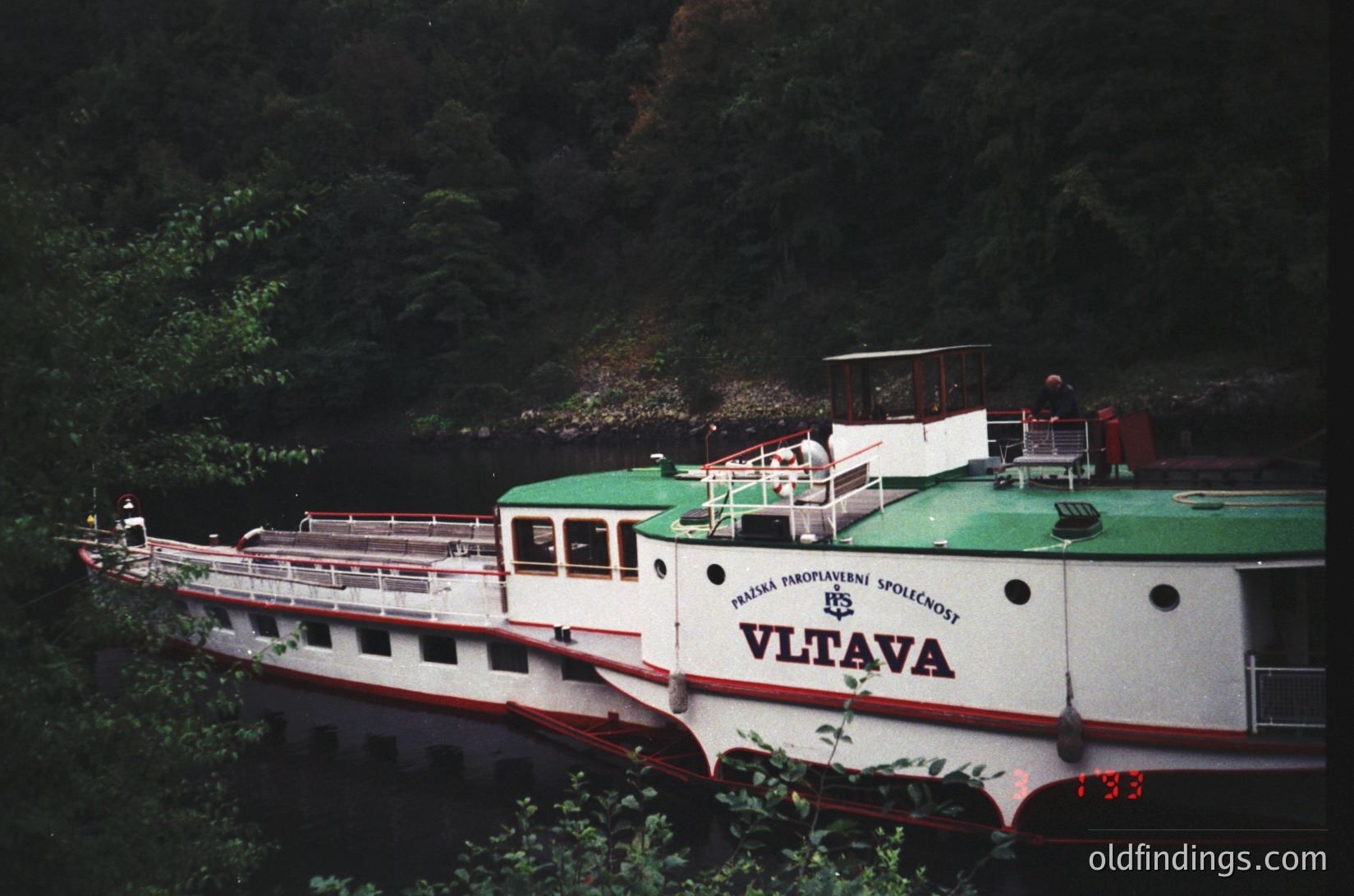 Vintage riverboat "Vltava" with green roof and white hull, moored along a forested riverbank. Czech text suggests connection to Prague’s historic steamship company (1960s-70s). Ideal for retro travel, industrial design, or Central European history research.