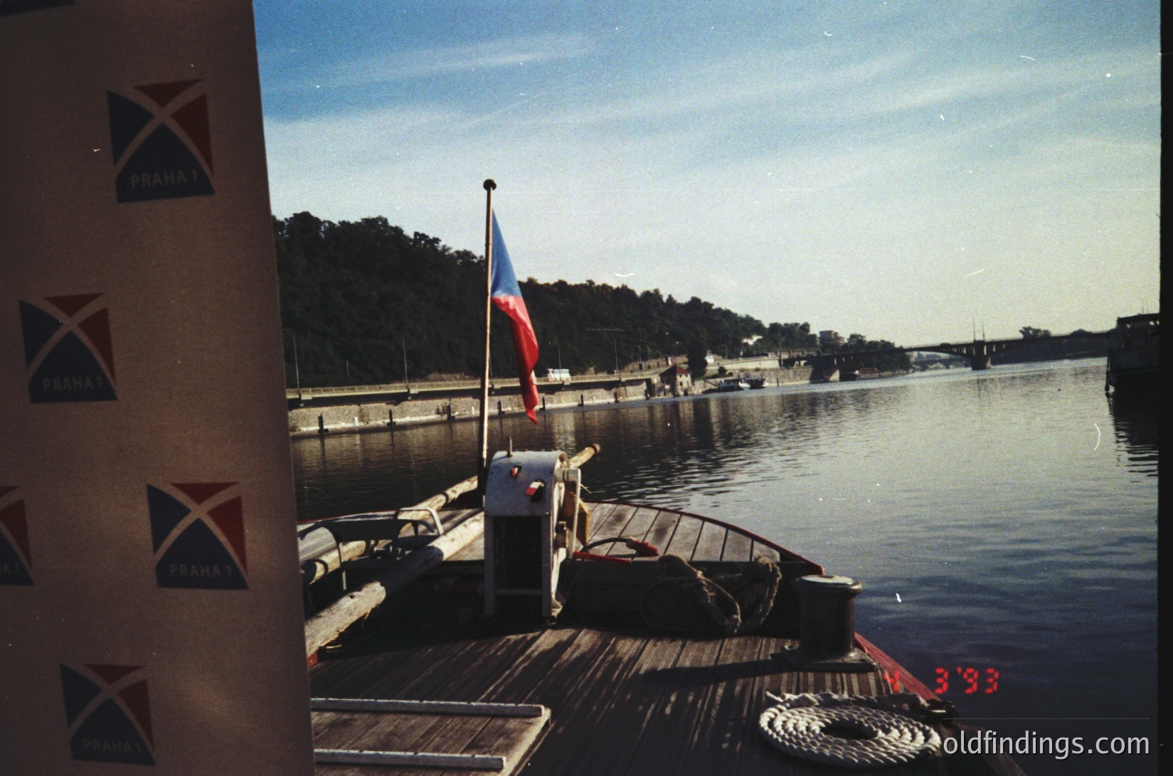 Vintage boat docked at Prague’s Vltava River, 1993. Czech flag flying, "PRAHA" branding on wooden pier. Urban greenery and historic architecture in background.