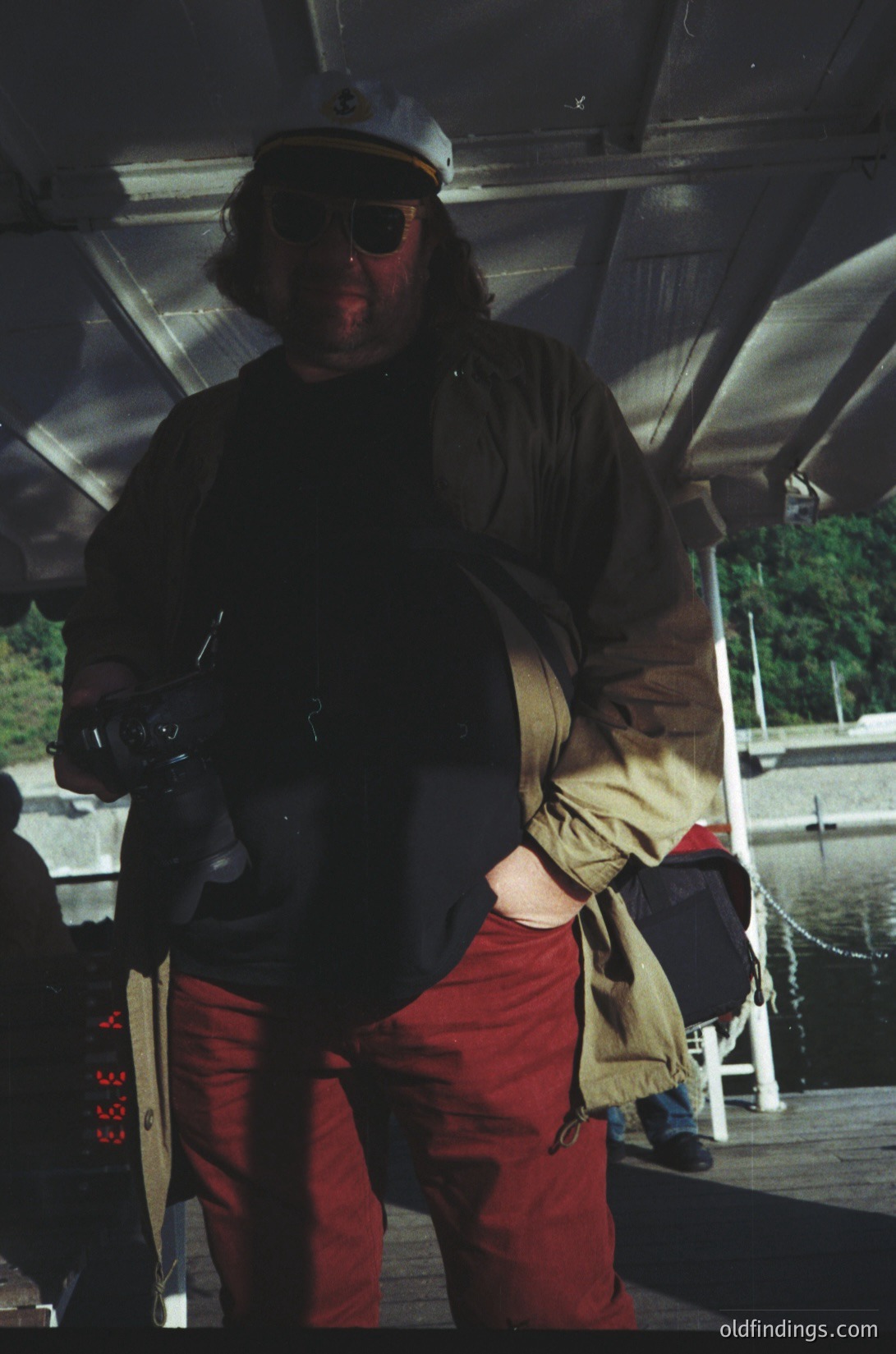 Vintage photo of a man in a naval officer’s peaked cap, sunglasses, and red trousers aboard a ship. Dark coat, beige jacket, and a camera strap suggest a maritime photographer or officer. Industrial metal hull and ropes in background. Likely mid-20th century naval or port setting.