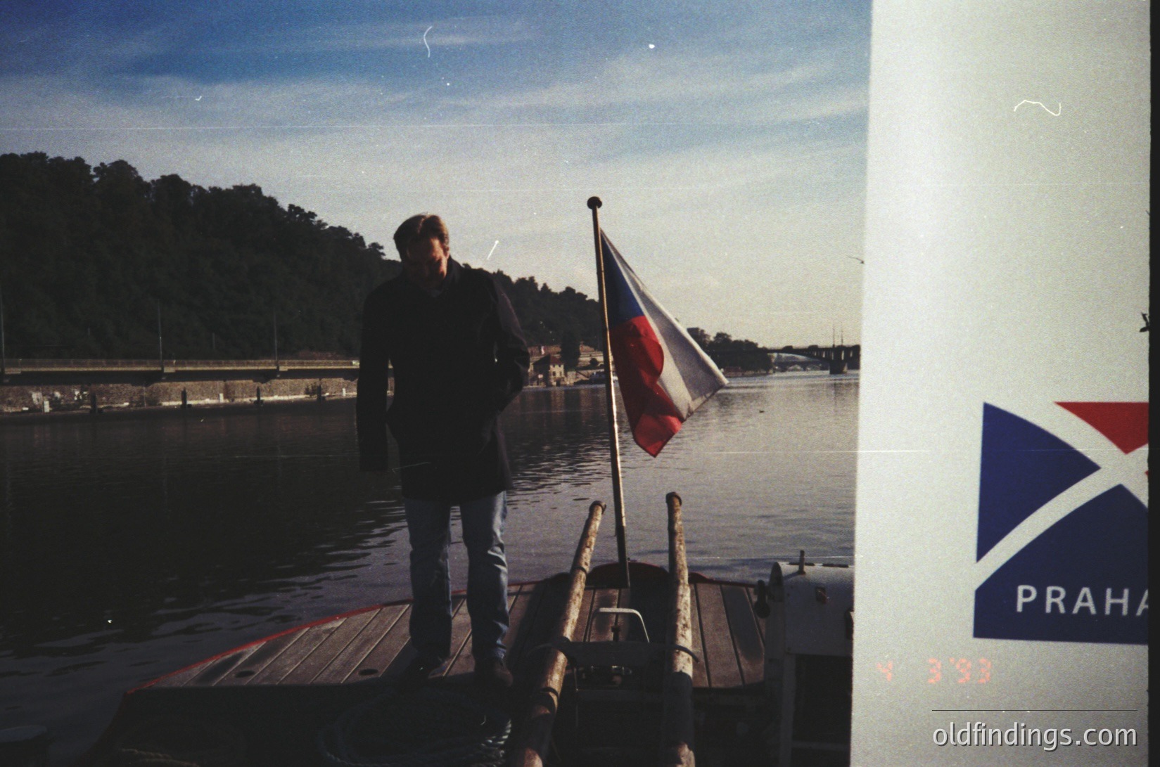 Vintage boat docked at waterfront with individual in dark jacket and bell-bottoms, holding a flag featuring red, white, and blue diagonal stripes. "PRAHA" logo visible on a wooden dock, suggesting a Czech connection. Trees and buildings in background indicate a European riverside or marina setting, likely 1970s–1980s.