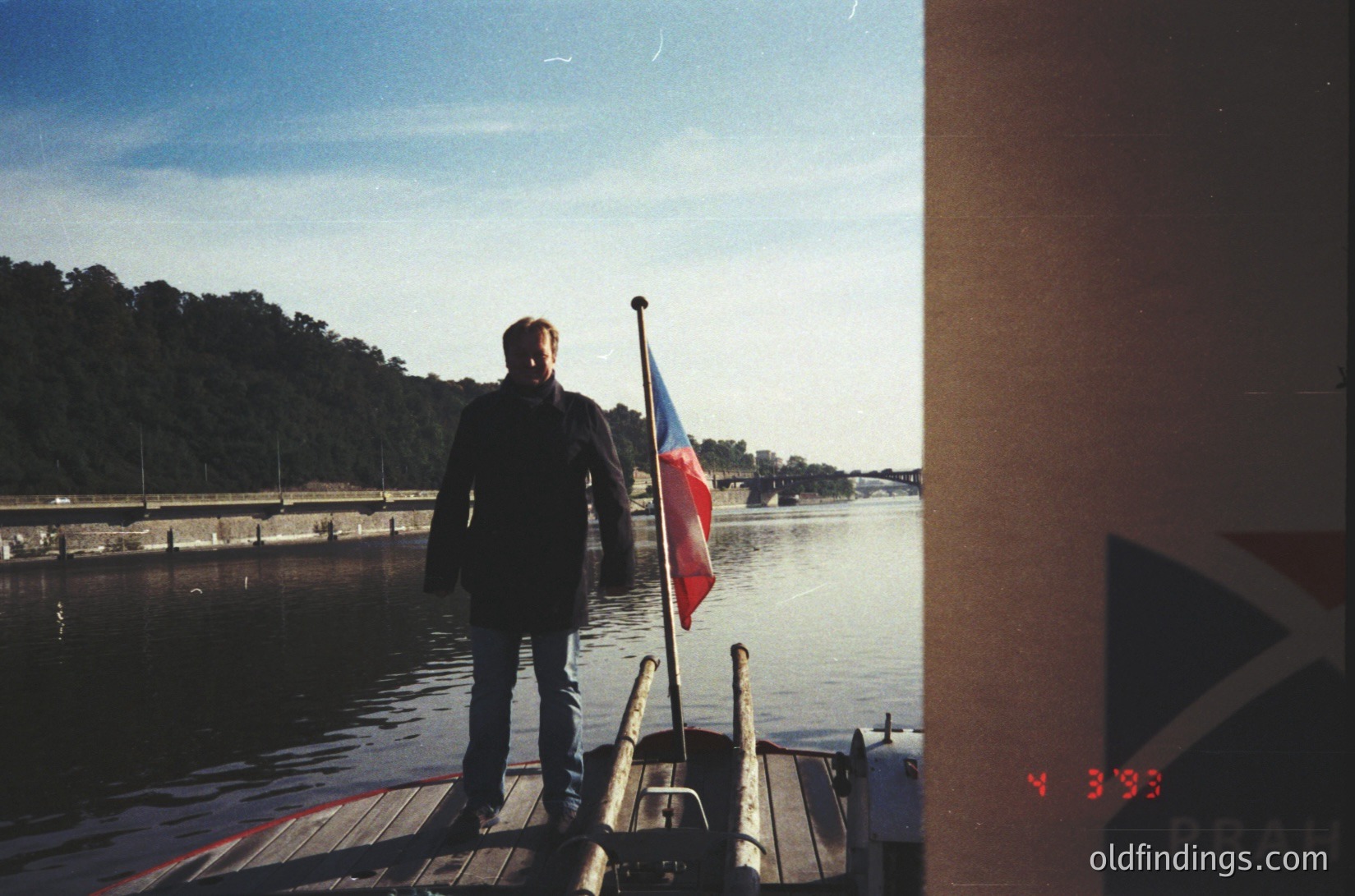 Vintage photo of a man standing on a wooden dock, holding a flagpole with the Czech Republic flag. Industrial waterfront with distant trees and a bridge. Overlay text reads "PRAHA 4 393," suggesting Prague, likely from the late 1980s or early 1990s.
