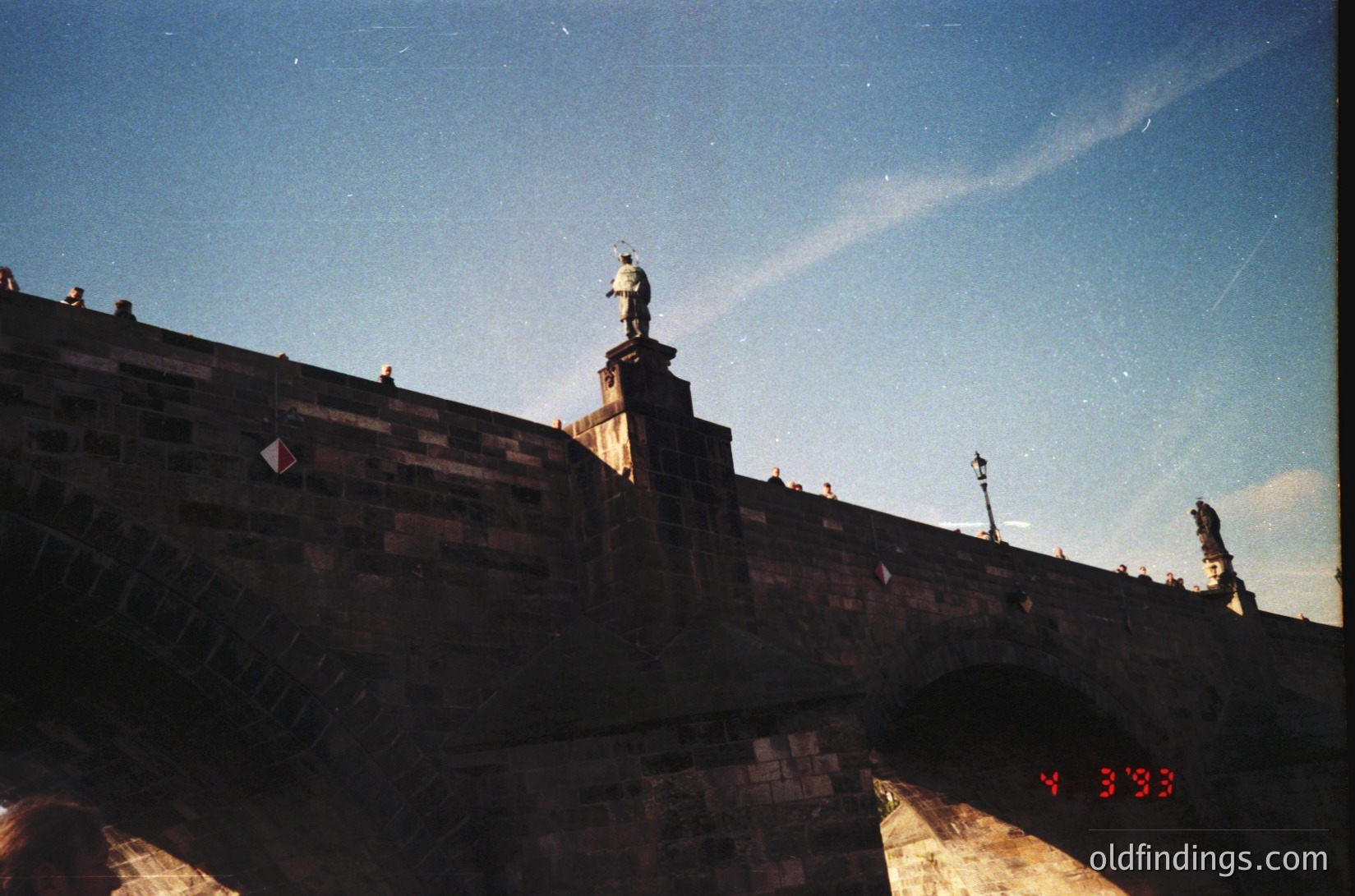 Vintage black-and-white shot of Prague’s Charles Bridge, featuring a statue atop a stone balustrade. Gothic architecture with medieval stonework and a lone figure in period attire. Overcast sky enhances dramatic lighting. Likely 1960s–1980s.