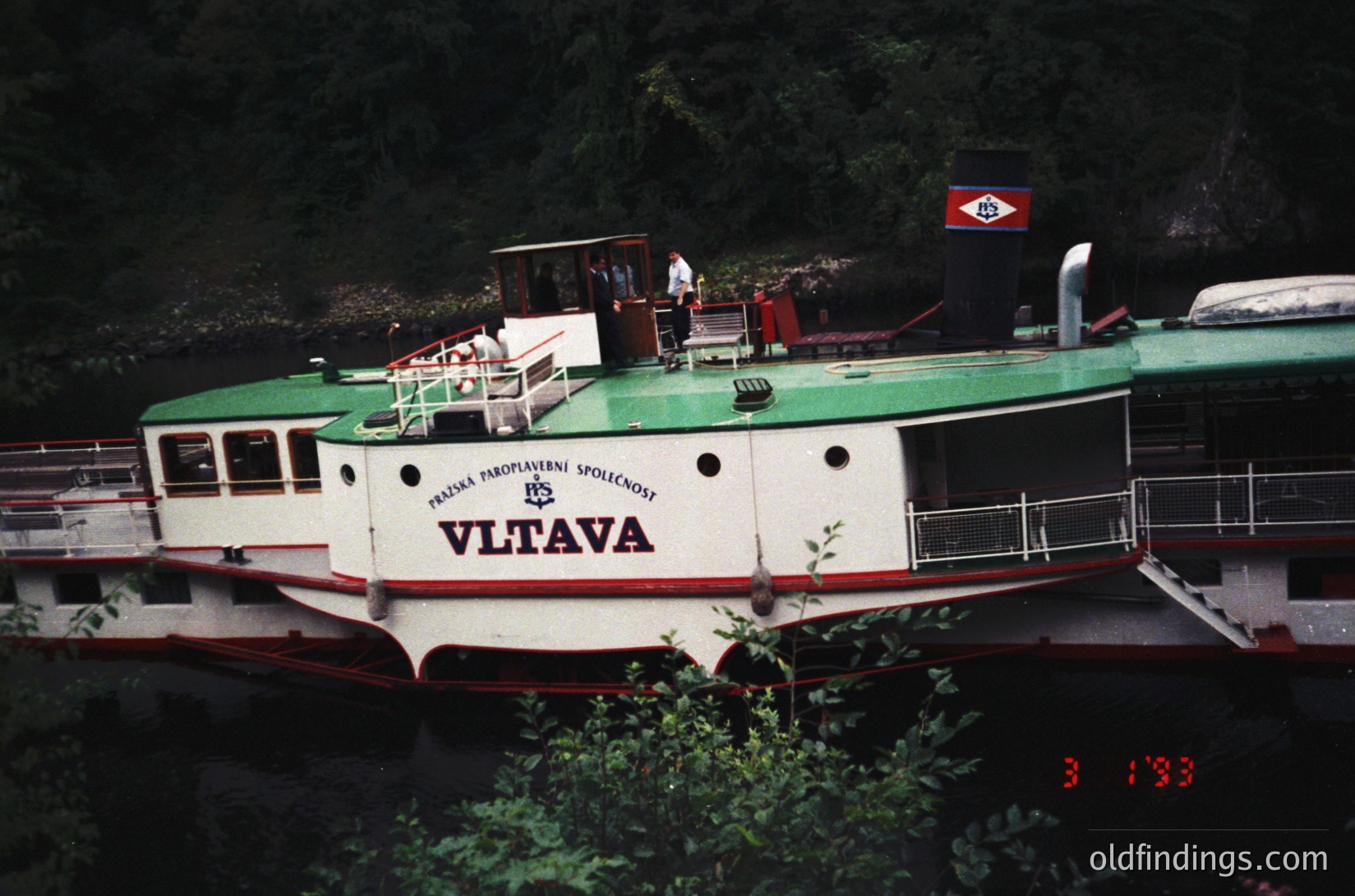 Vintage riverboat "Vltava" with green and white livery, reflecting mid-20th century Czech design. Prominent "Praha" (Prague) maritime society logo on hull. Wooded riverbank and calm water in background. Likely used for passenger transport or tourism.