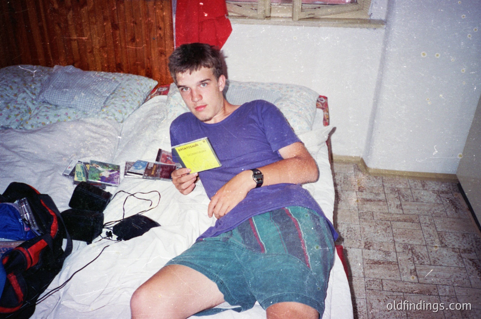 Young man in 1990s-era casual wear (purple T-shirt, cargo shorts) poses indoors with a vintage Nintendo Game Boy and yellow manual. Surrounding items include a black backpack, CDs, and a tiled floor. Likely late 1990s/early 2000s.